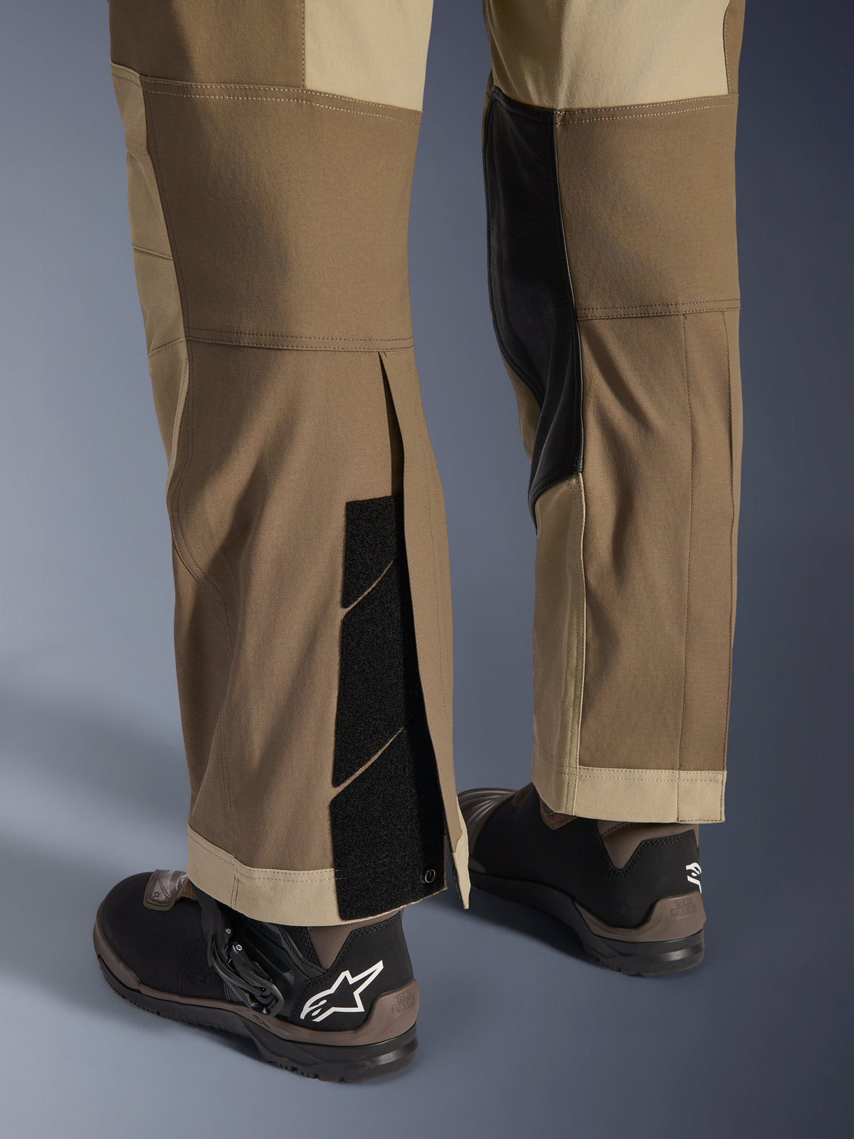 Calças Alpinestars Maxdura Dual em Walnut Aluminum, vista detalhada da parte inferior da perna e da bainha com design por cima da bota, fechos de velcro ajustáveis e painéis interiores reforçados, combinadas com botas de motociclismo pretas.