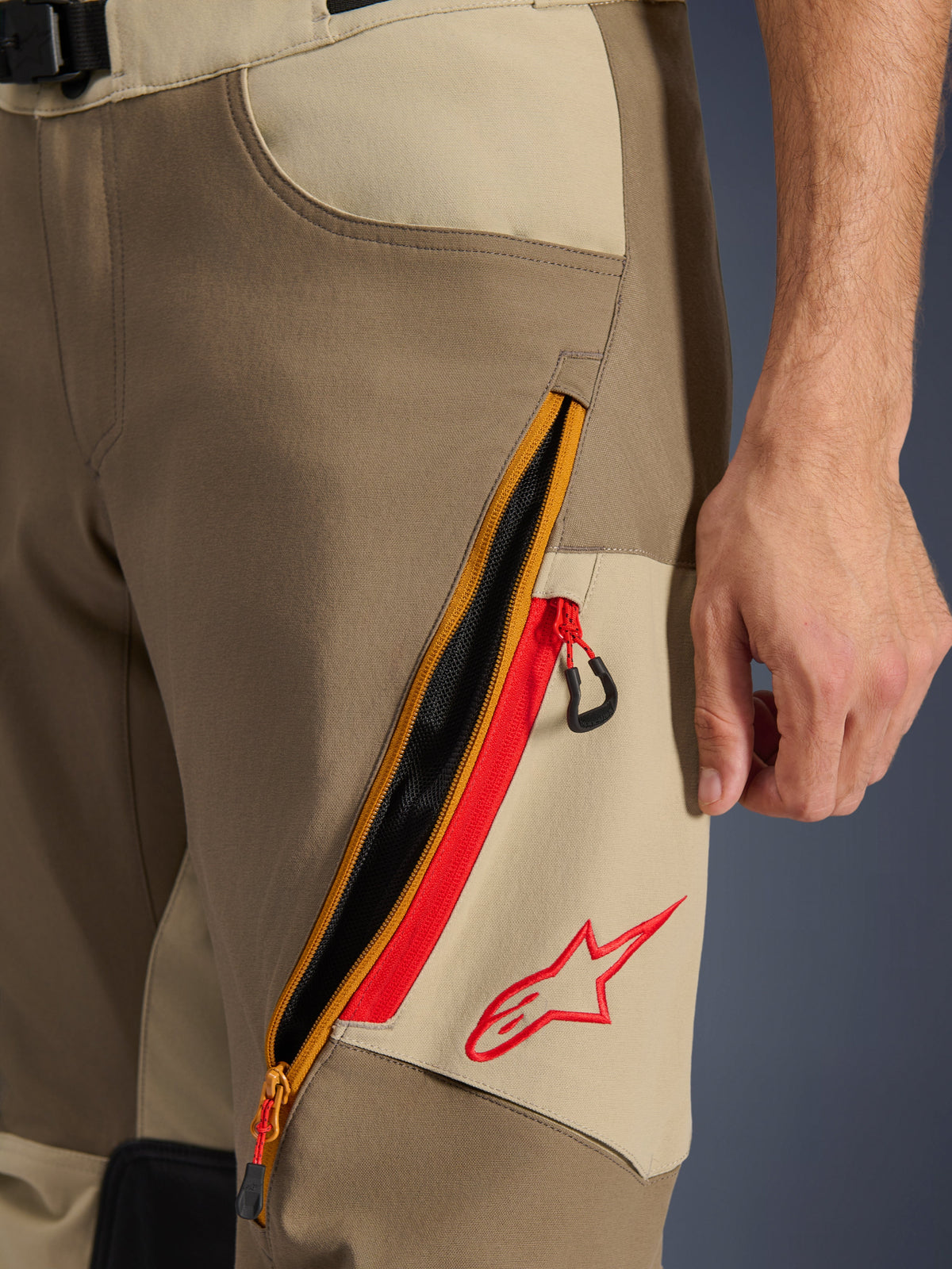 Alpinestars Maxdura Dual Pants, Walnut e Aluminum, pormenor da coxa com um grande sistema de ventilação diagonal com fecho de correr e detalhes vermelhos, logótipo Alpinestars vermelho num painel bege, equipamento de motociclismo de aventura