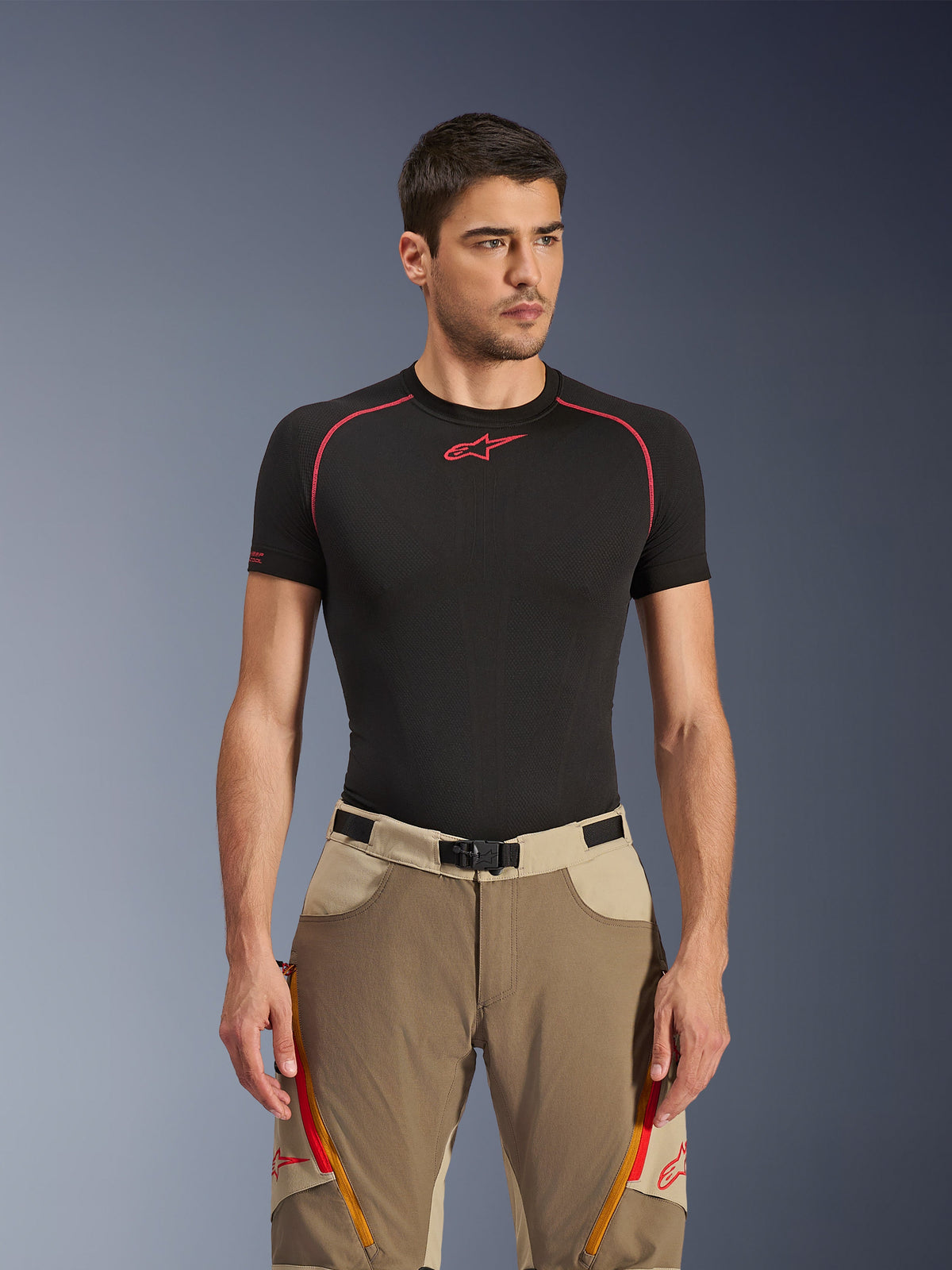 Alpinestars Maxdura Dual Pants em Walnut Aluminum, calças de motociclismo de aventura beges e castanhas, vestidas por um modelo com uma camisola interior preta, com aberturas de ventilação cor de laranja com fecho nas coxas, logótipos vermelhos e fivela magnética na cintura, retrato de estúdio.