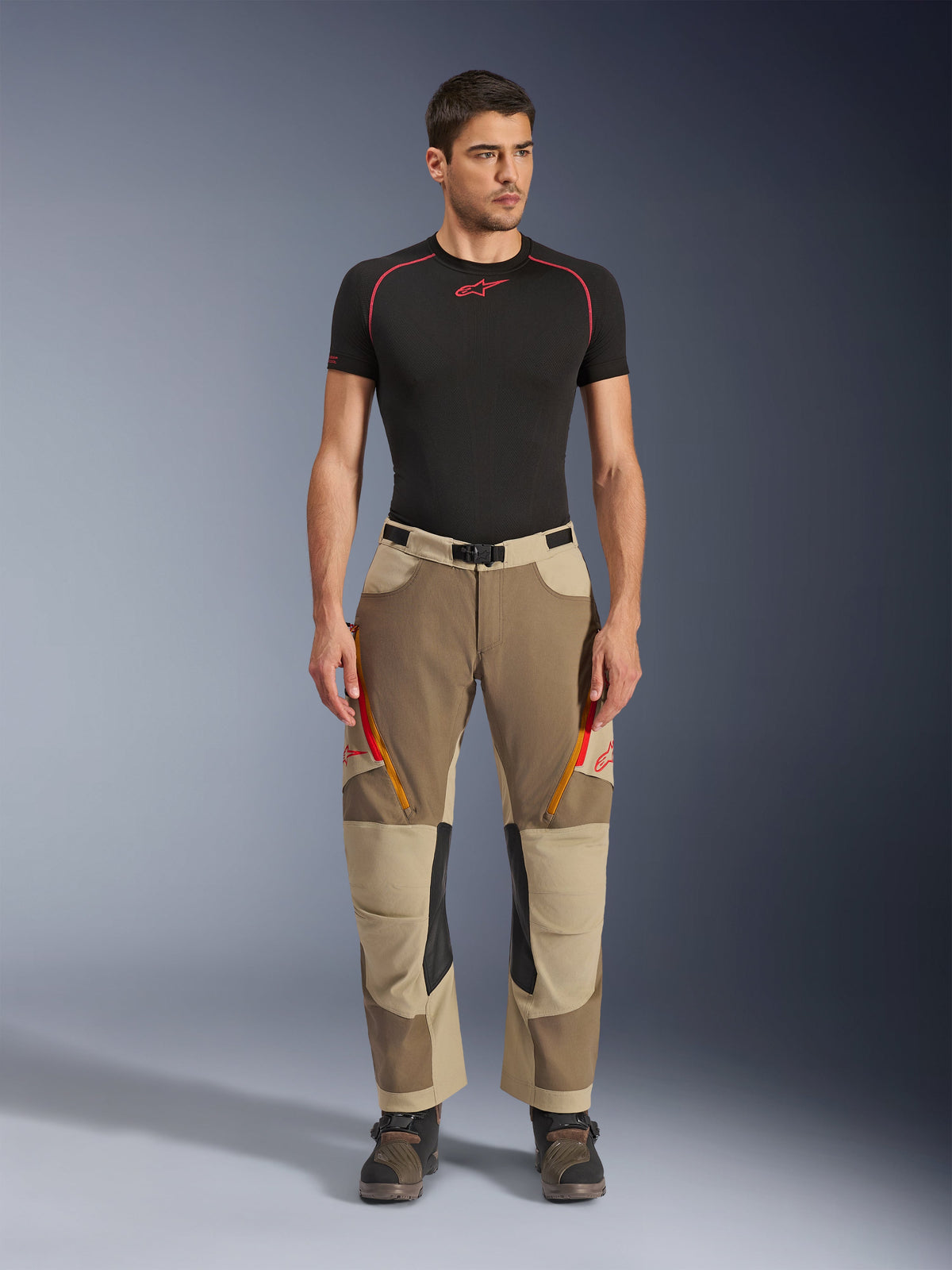 Alpinestars Maxdura Dual Pants, calças de aventura e dual-sport, cor Walnut Aluminum, usadas com uma camisola de compressão preta de manga curta e botas de aventura, com aberturas de ventilação cor-de-laranja com fecho e logótipos vermelhos nas coxas, cinto preto com fivela e design para usar por cima da bota para condução de motociclos.