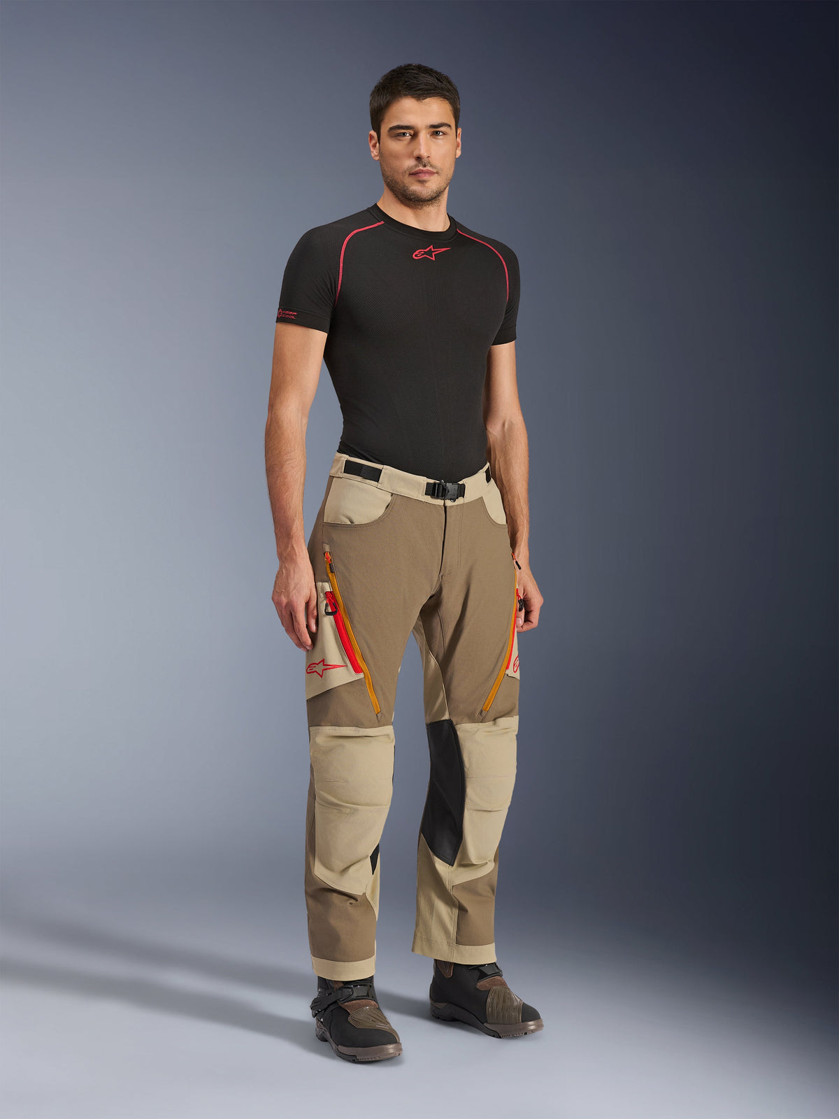 Alpinestars Maxdura Dual Pants em Walnut Aluminum, calças de motociclismo de aventura em castanho e bege com fechos de correr cor-de-laranja e painéis nos joelhos em pele preta, usadas com uma t-shirt de camada de base preta e botas de aventura, vista frontal de corpo inteiro