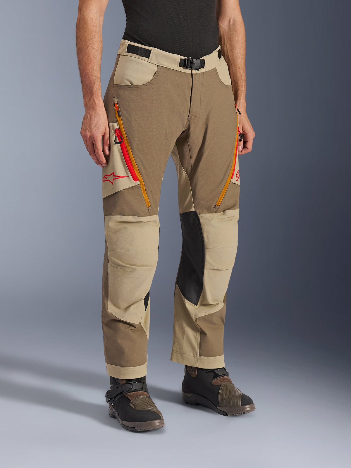 Calças Alpinestars Maxdura Dual, calças de aventura em castanho Walnut Aluminum e caqui, com aberturas de ventilação nas coxas com fecho em laranja e vermelho, painéis interiores dos joelhos em pele preta reforçada e logótipo Astars vermelho, usadas com botas técnicas