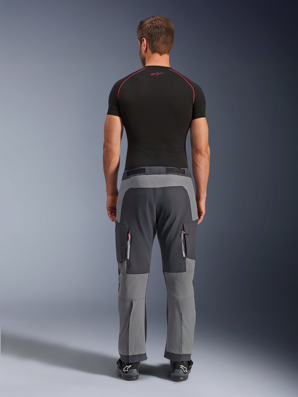 Alpinestars Maxdura Dual Pants, calças de motociclismo de aventura em cinzento escuro e cinzento profundo, vista traseira num modelo a usar t-shirt preta e botas, com painéis cinzentos de vários tons e bolsos cargo com fecho e detalhes vermelhos.