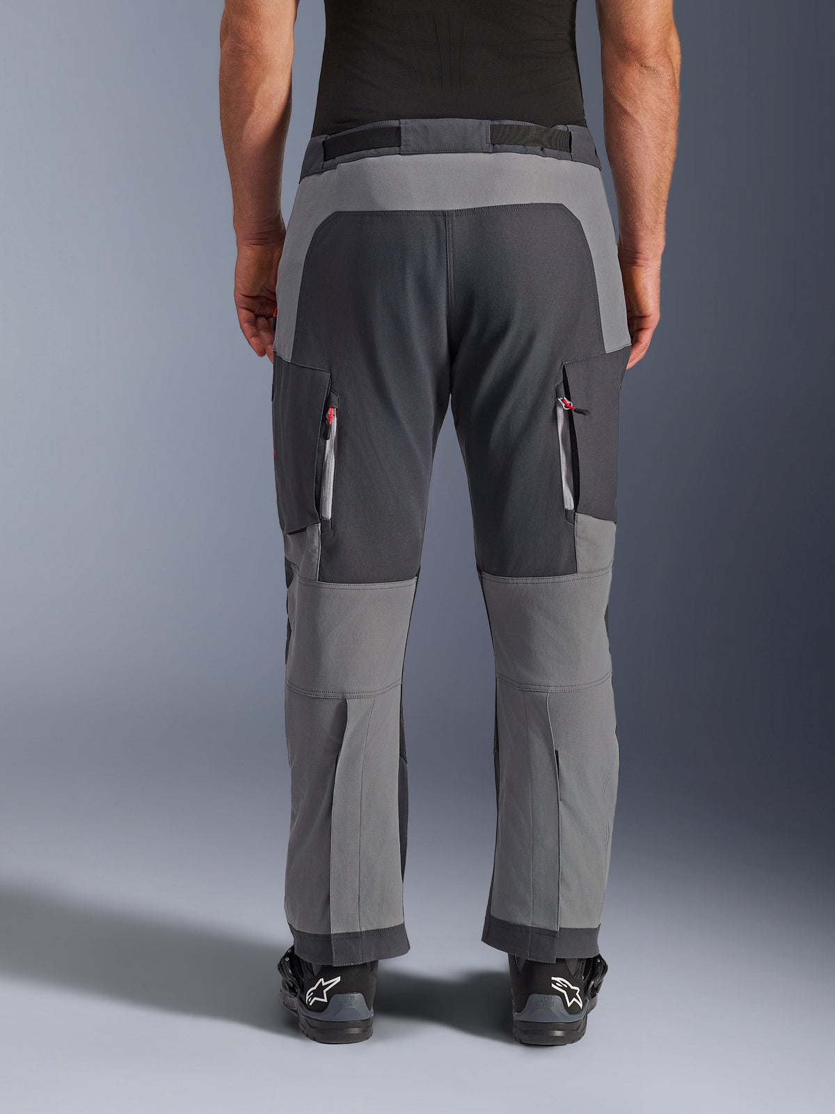 Alpinestars Maxdura Dual Pants, calças de motociclismo de aventura, cinzento escuro e cinzento profundo, vista traseira com destaque para os painéis reforçados no assento e joelhos, aberturas de ventilação com fecho de correr nas coxas, usadas com botas Alpinestars pretas, construção robusta em poliamida elástica para condução dual-sport
