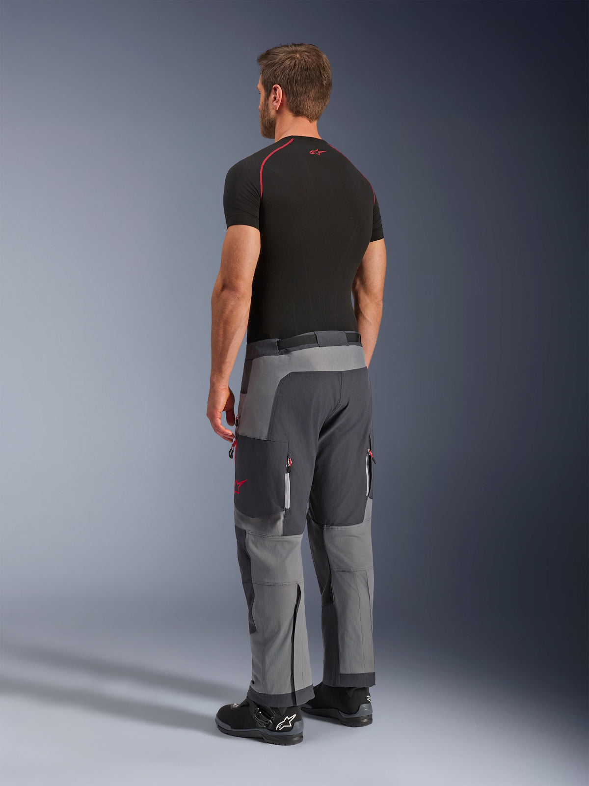 Alpinestars Maxdura Dual Pants, calças de motociclismo de aventura, cinzento escuro e cinzento profundo, vestidas por um modelo numa vista lateral traseira, com detalhes vermelhos nos fechos e no logótipo, painéis cinzentos reforçados, bolsos cargo e ajuste sobre a bota