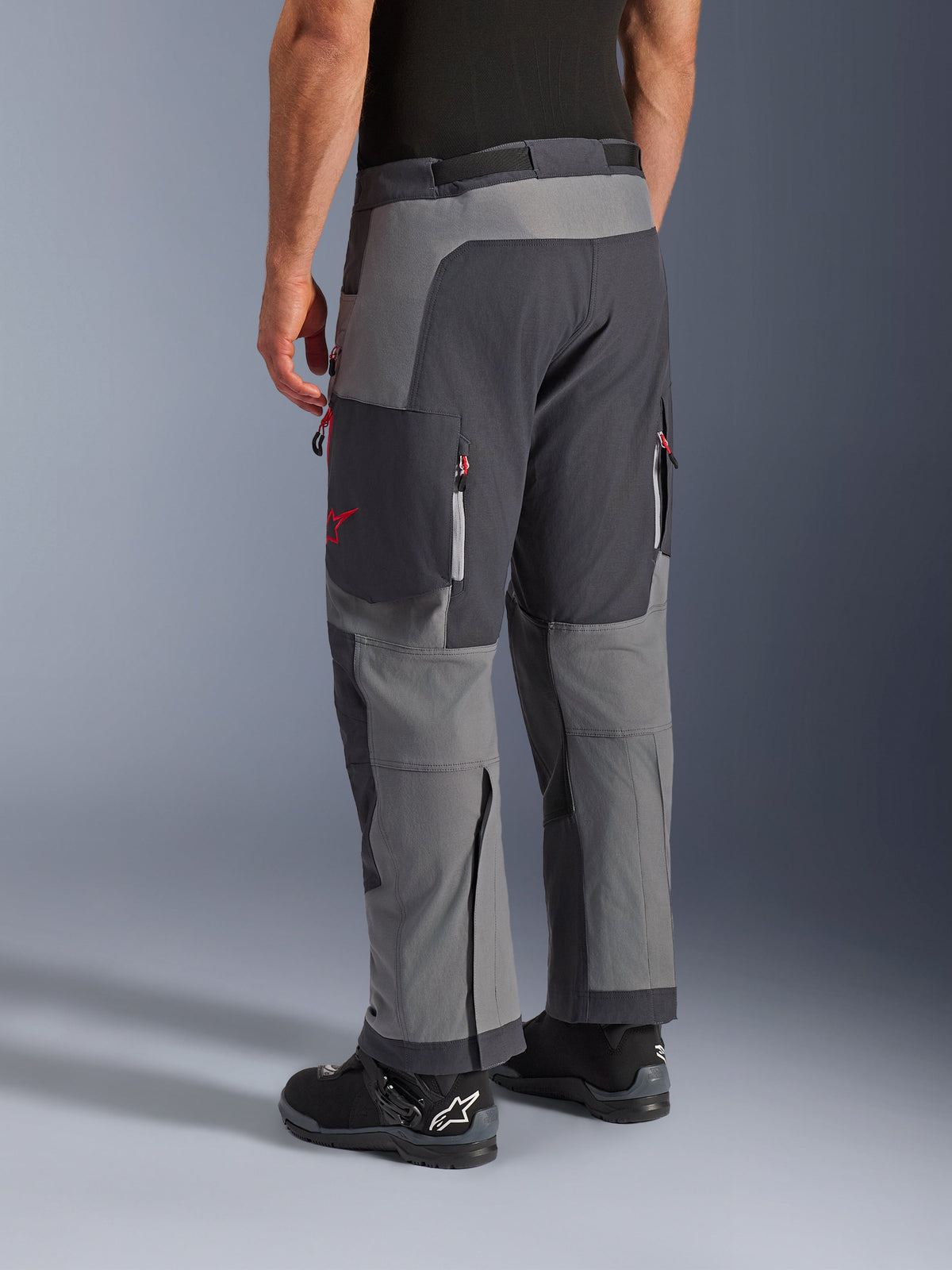 Alpinestars Maxdura Dual Pants, calças de motociclismo de aventura, cinzento escuro e cinzento profundo, vista traseira com construção multi-painel, detalhes de fechos vermelhos e logótipo de estrela, combinadas com botas Alpinestars pretas, concebidas para performance dual-sport