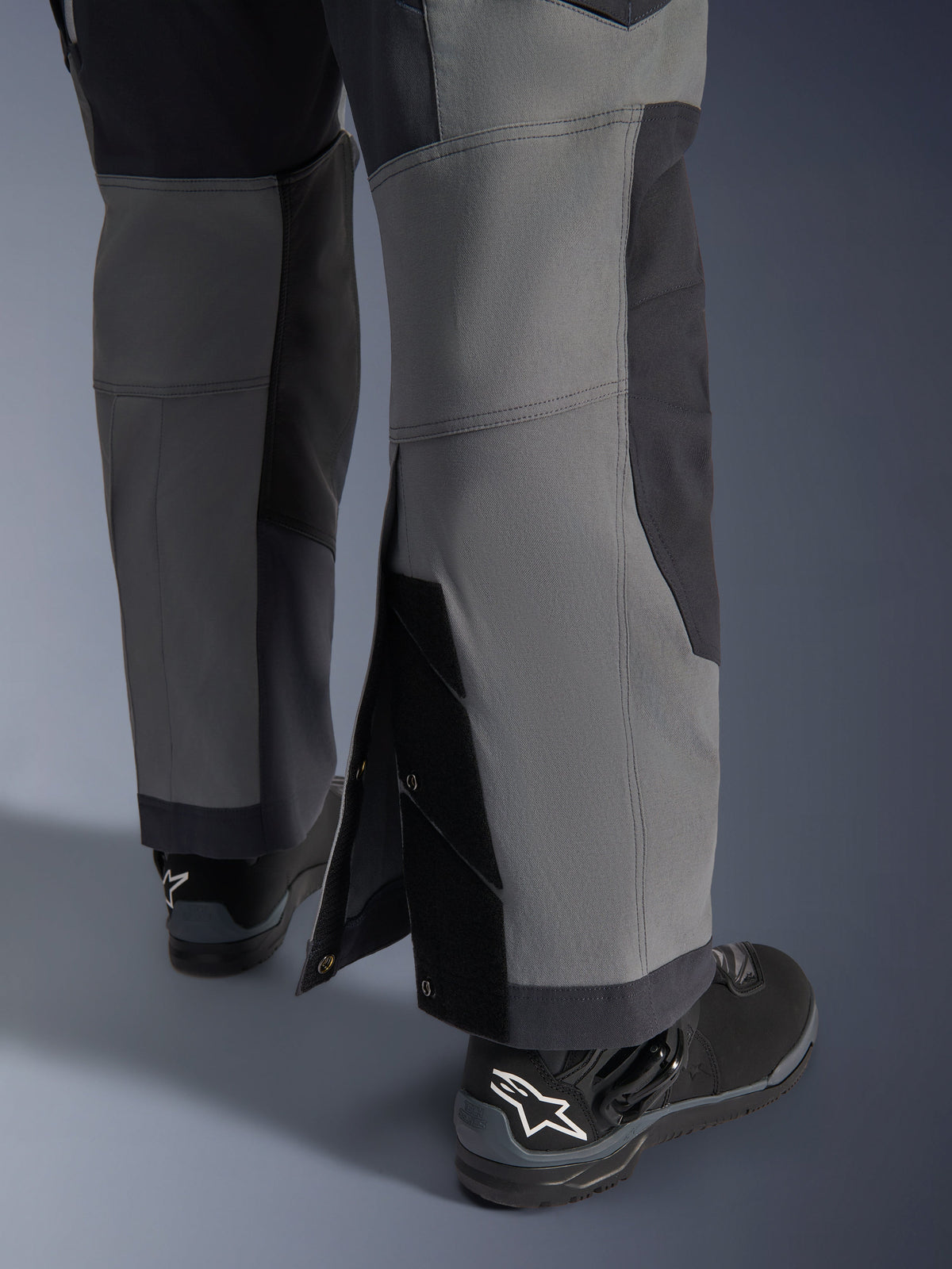 Alpinestars Maxdura Dual Pants, calças de motociclismo de aventura, cinzento escuro e cinzento profundo, vista detalhada da bainha ajustável sobre a bota com fechos de mola e painéis reforçados, usadas com botas Alpinestars pretas, design técnico robusto para condução dual-sport