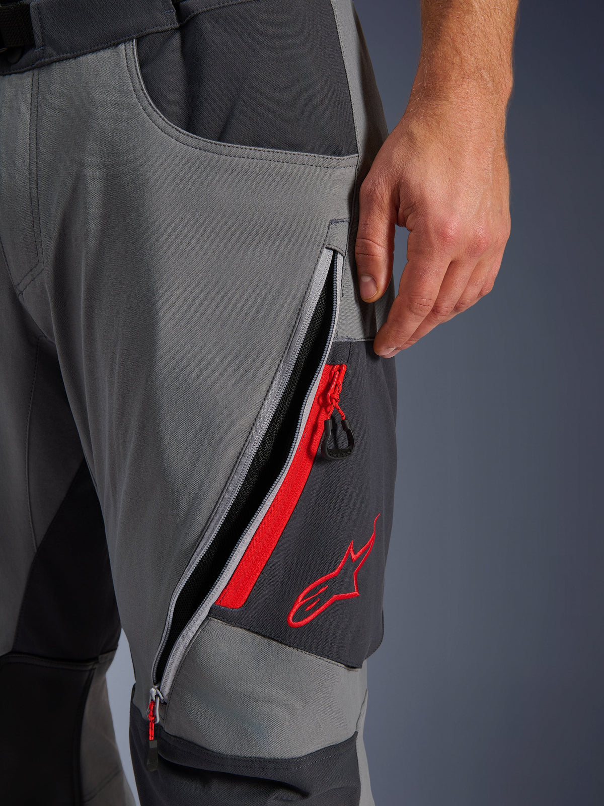 Alpinestars Maxdura Dual Pants, calças de motociclismo de aventura em cinzento escuro e cinzento profundo, vista em pormenor da abertura de ventilação na coxa com fecho de correr e detalhes em vermelho, logótipo de estrela bordado a vermelho, design em tecido elástico técnico