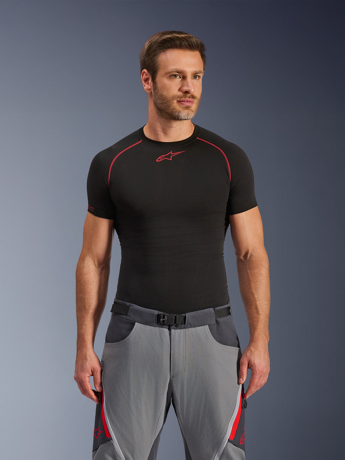 Calças Alpinestars Maxdura Dual, cinzento escuro e cinzento profundo com detalhes vermelhos, usadas com uma camisola base layer preta com costuras vermelhas, corte atlético com cinto de fivela magnética integrado, equipamento de motociclismo de aventura durável