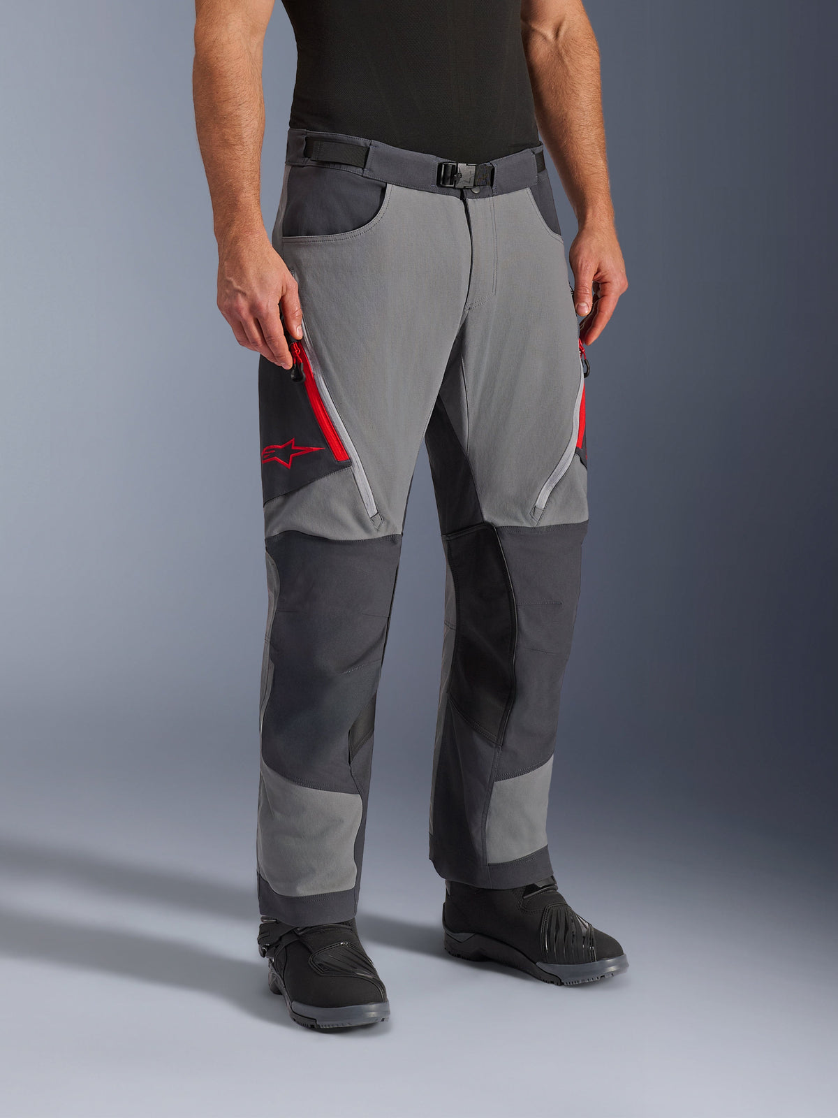 Alpinestars Maxdura Dual Pants em Cinzento Escuro e Cinzento Profundo, calças de motociclismo adventure-touring com aberturas de ventilação nas coxas com fecho de correr vermelho e logótipo de estrela, painéis escuros reforçados nos joelhos, combinadas com botas de motociclismo pretas