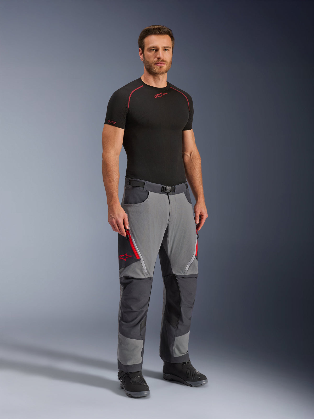 Alpinestars Maxdura Dual Pants, calças de motociclismo de aventura, cinzento escuro e cinzento profundo, usadas com uma camada base preta e botas, com aberturas de ventilação vermelhas com fecho de correr, painéis reforçados nos joelhos e assento, e cintura com cinto, corte atlético para condução dual-sport