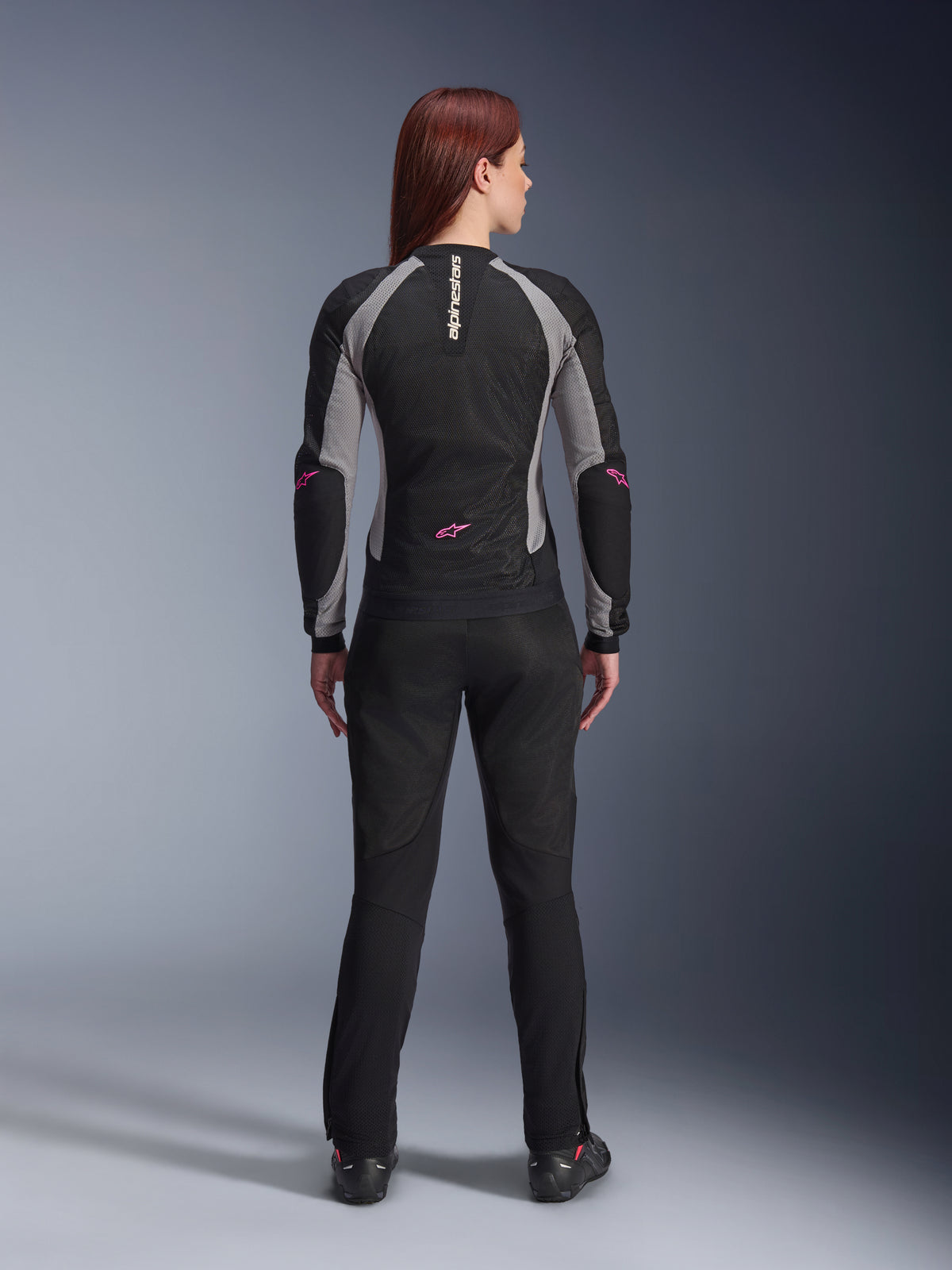 Calças Alpinestars Stella Troop-Air para Mulher em preto, vista traseira, combinadas com um casaco de motociclismo em malha preto e cinzento com detalhes cor-de-rosa, fotografia de estúdio destacando o design têxtil técnico ventilado