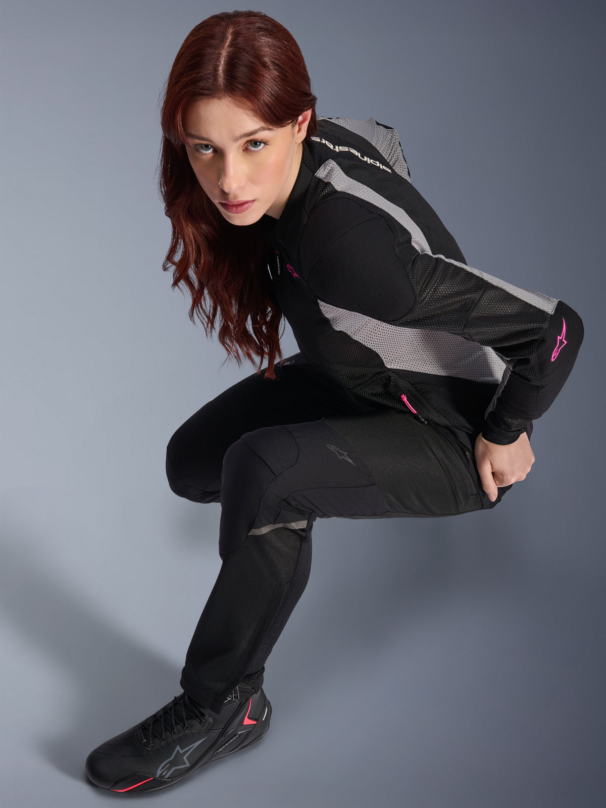Mulher a usar calças Alpinestars Stella Troop-Air pretas, calças têxteis leves e ventiladas, combinadas com um casaco de malha preto e cinzento com detalhes rosa e botas de motociclismo pretas, retrato de estúdio, equipamento de condução urbana