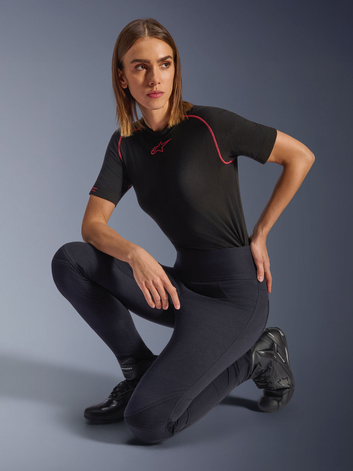 Leggings Stella Flex-Ast da Alpinestars, pretas, usadas por uma mulher ajoelhada em estúdio, combinadas com um top de performance preto de manga curta com detalhes cor-de-rosa e botas de motociclismo pretas, com cintura alta e painéis flexíveis para condução urbana.