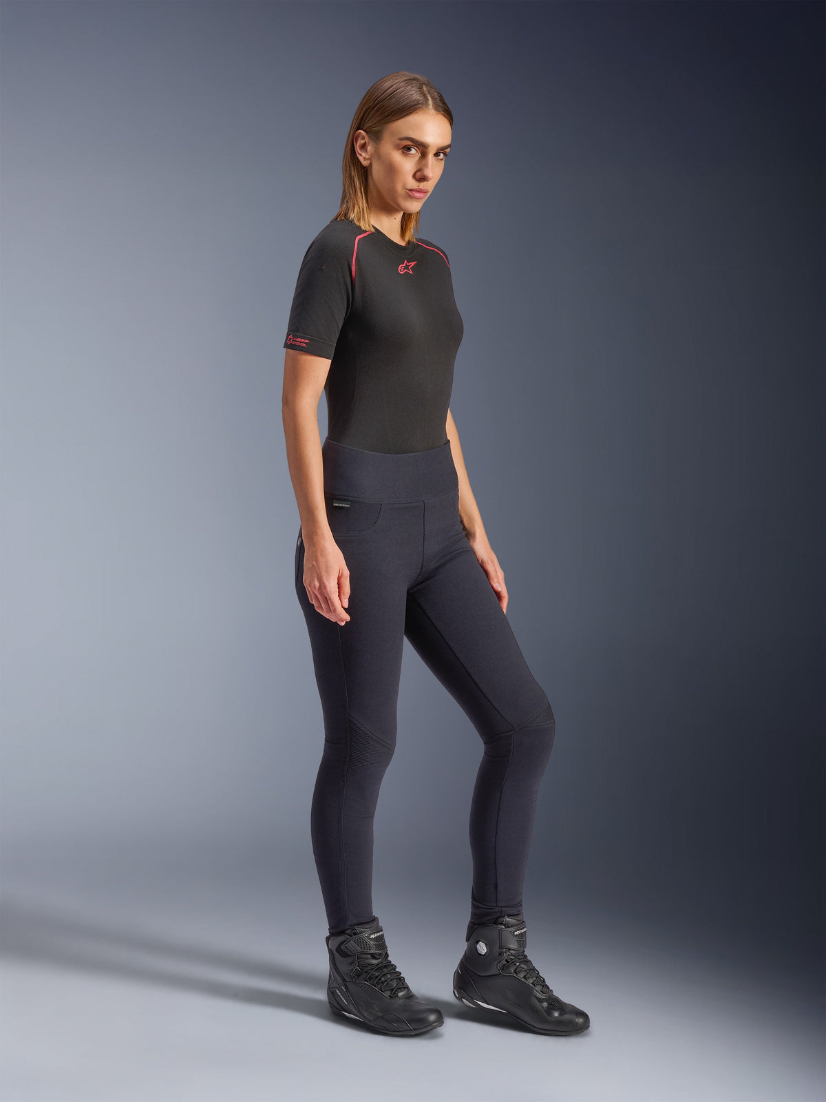 Alpinestars Stella Flex-Ast Leggings, leggings de motociclismo para mulher em preto, design de cintura alta e corte justo, usadas com uma t-shirt preta com detalhes vermelhos e botas de motociclismo pretas, fotografia de estúdio de corpo inteiro