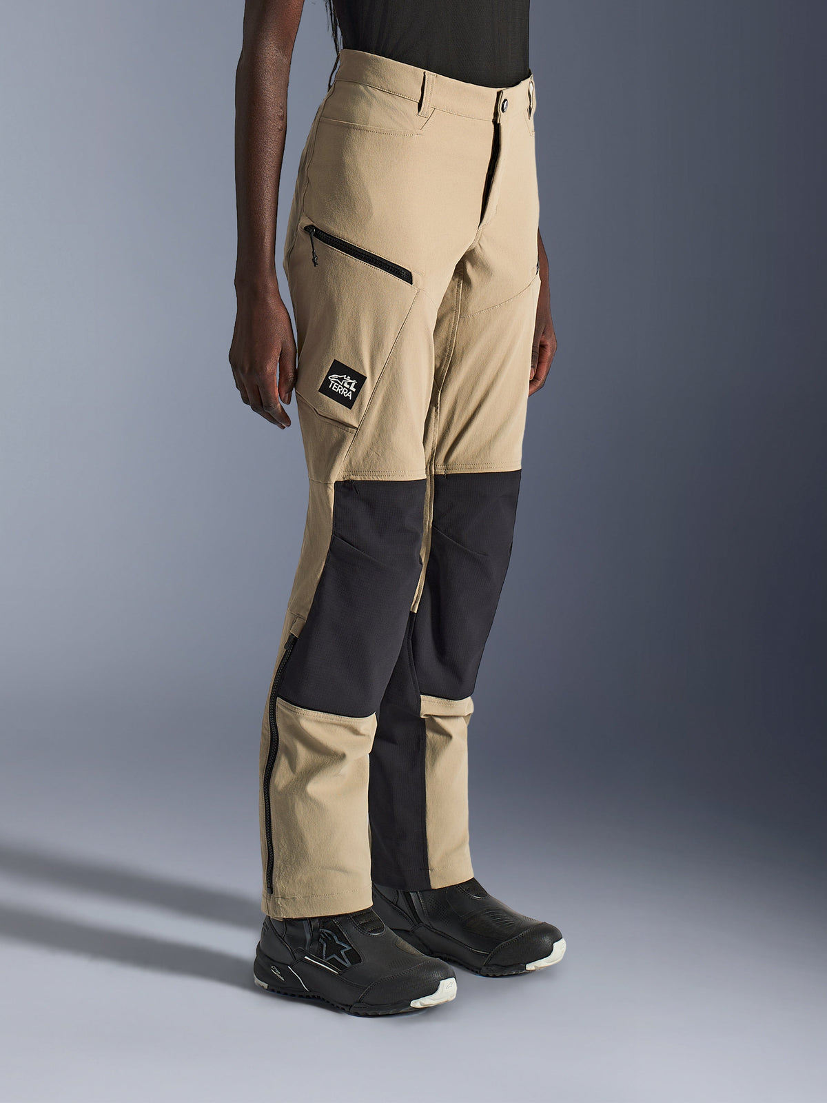 Woman wearing Alpinestars Stella Flex-AST Explorer Pants, Sand and Black adventure motorcycle pants with black ripstop knee panels, zippered cargo pockets, 'ALL TERRA' logo patch, and lower leg zippers.