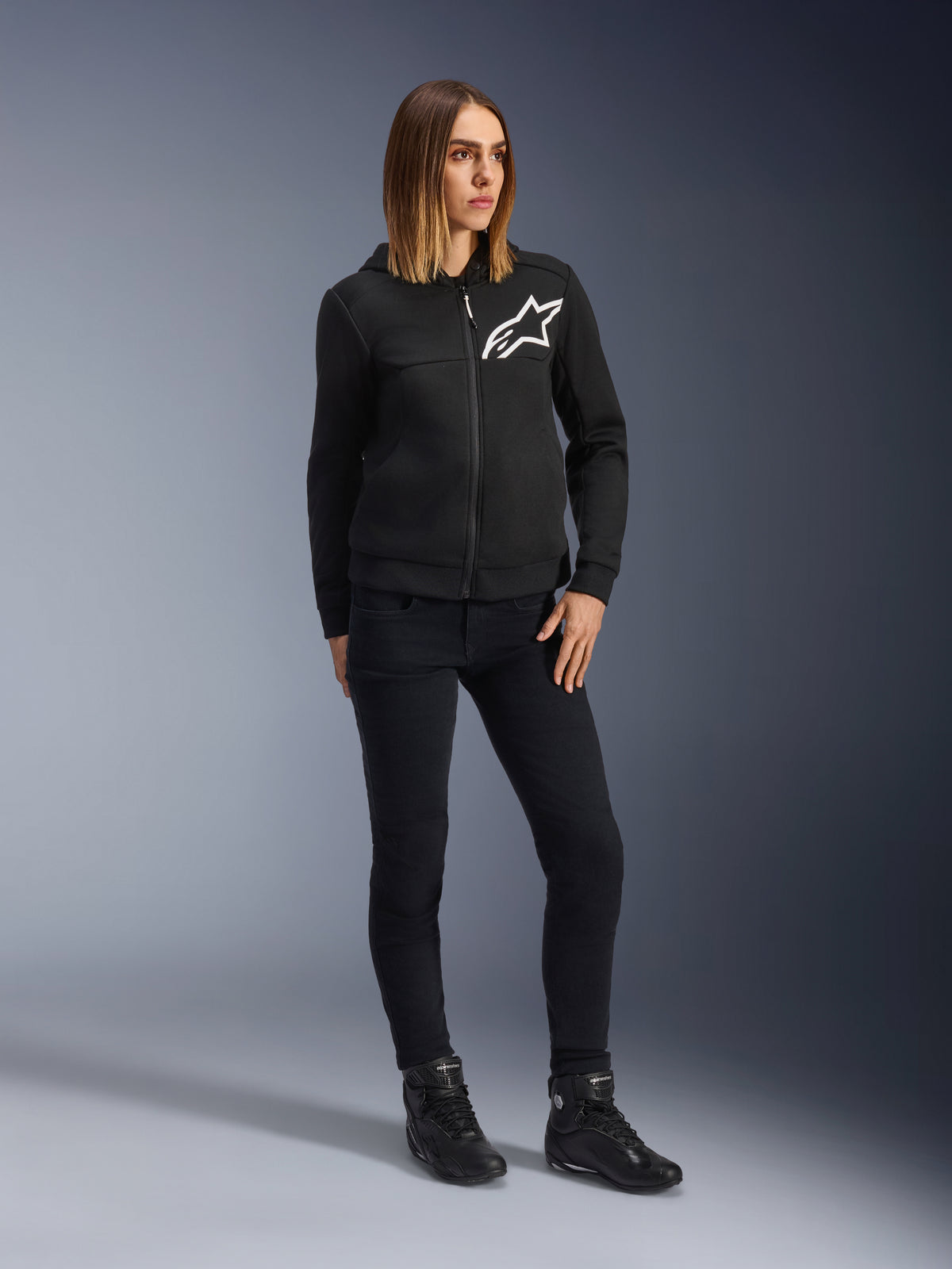 Mulher a usar Alpinestars Daisy V3 Women's Riding Denim, calças de ganga de motociclismo slim-fit em Rinse Black, combinadas com um hoodie preto com fecho e botas de motociclismo, equipamento de proteção urbano casual