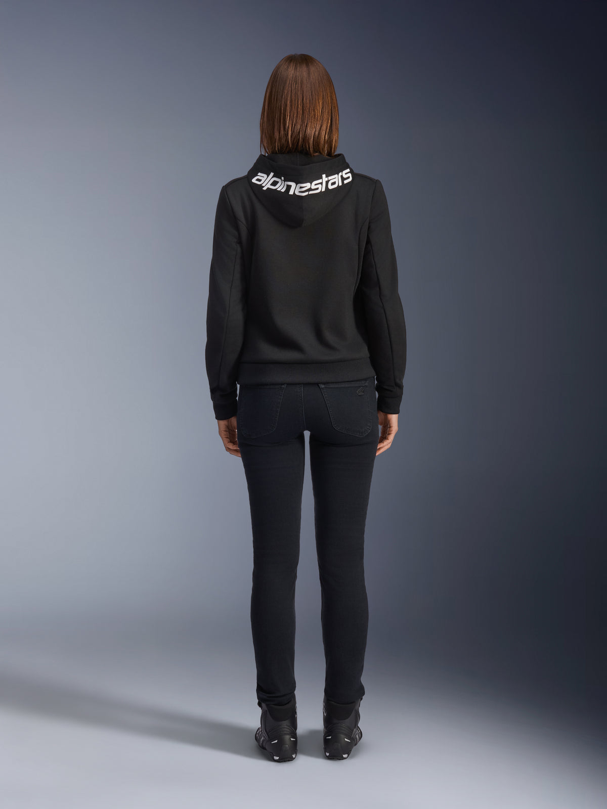 Calças de Ganga Daisy V3 Femininas Alpinestars, Black Rinse, calças de motociclismo de corte justo, vista traseira exibindo a cintura subida atrás e a silhueta feminina, combinadas com hoodie Alpinestars preto e botas de condução pretas, equipamento de condução urbano casual