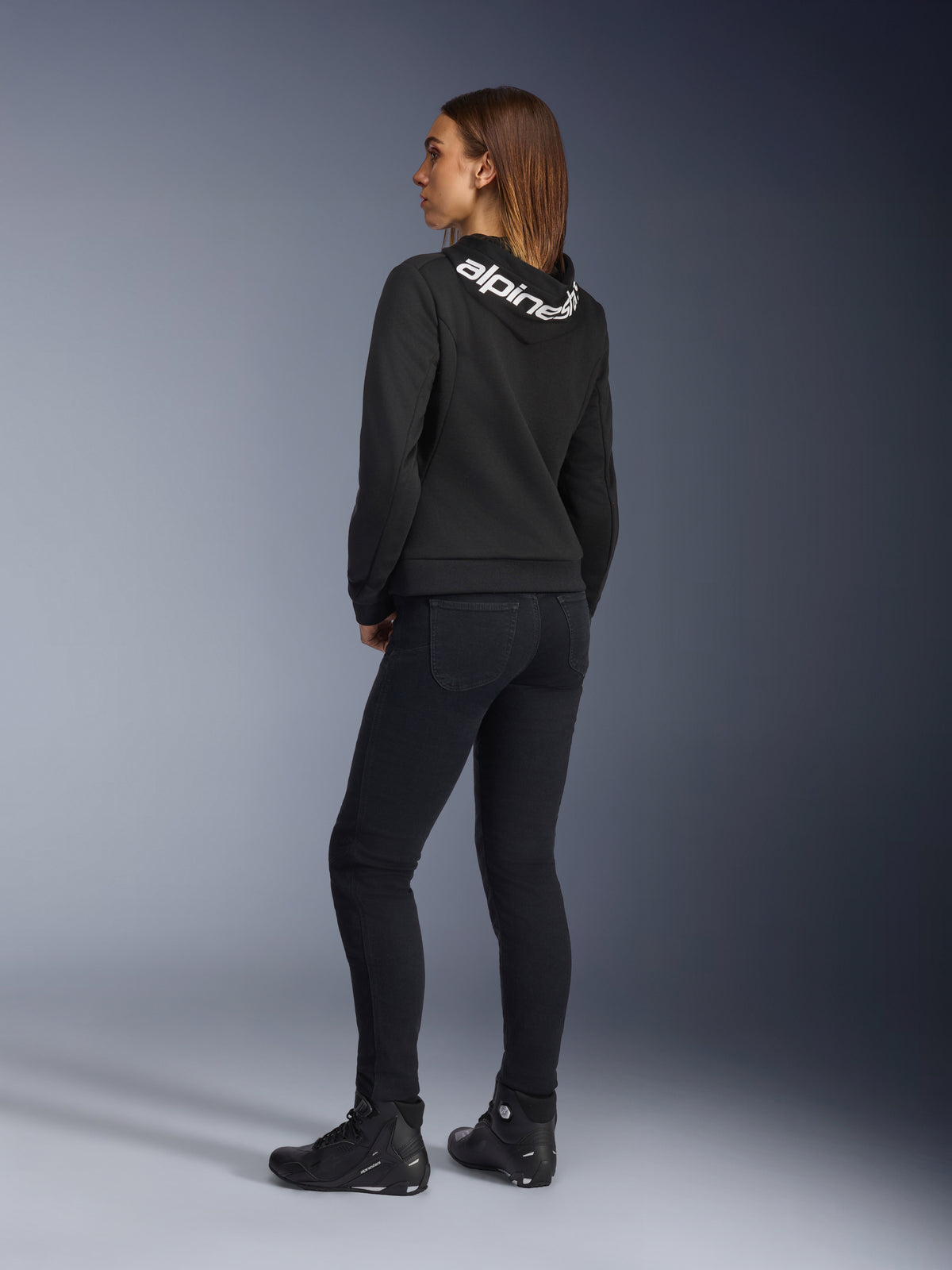 Alpinestars Daisy V3 Women's Riding Denim, calças de motociclismo para mulher, Rinse Black, vista traseira a mostrar o corte slim e a cintura subida, combinadas com um hoodie preto Alpinestars e botas de condução para um visual urbano de motociclista
