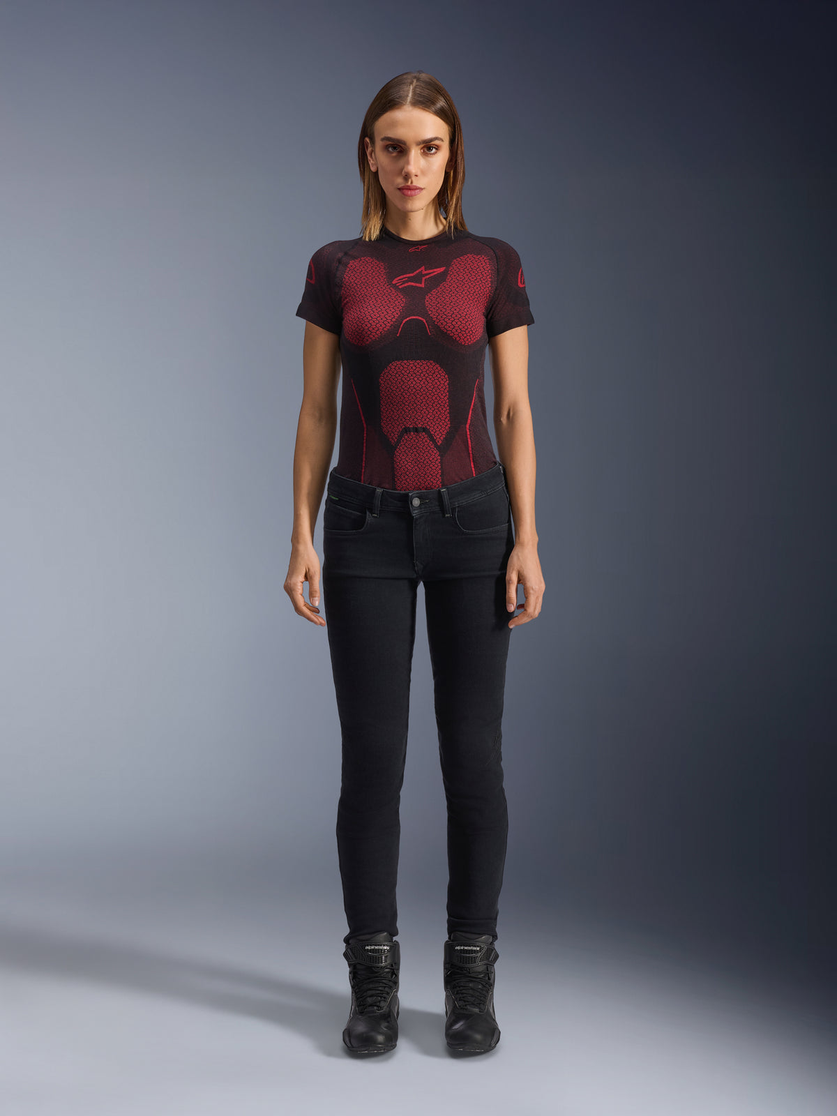 Alpinestars Daisy V3 Women's Riding Denim, Rinse Black, calças de motociclismo de corte justo, vestidas por uma modelo com uma t-shirt técnica com padrão vermelho e preto e botas de motociclismo pretas, equipamento de condução urbana casual com silhueta feminina e design de cintura subida.