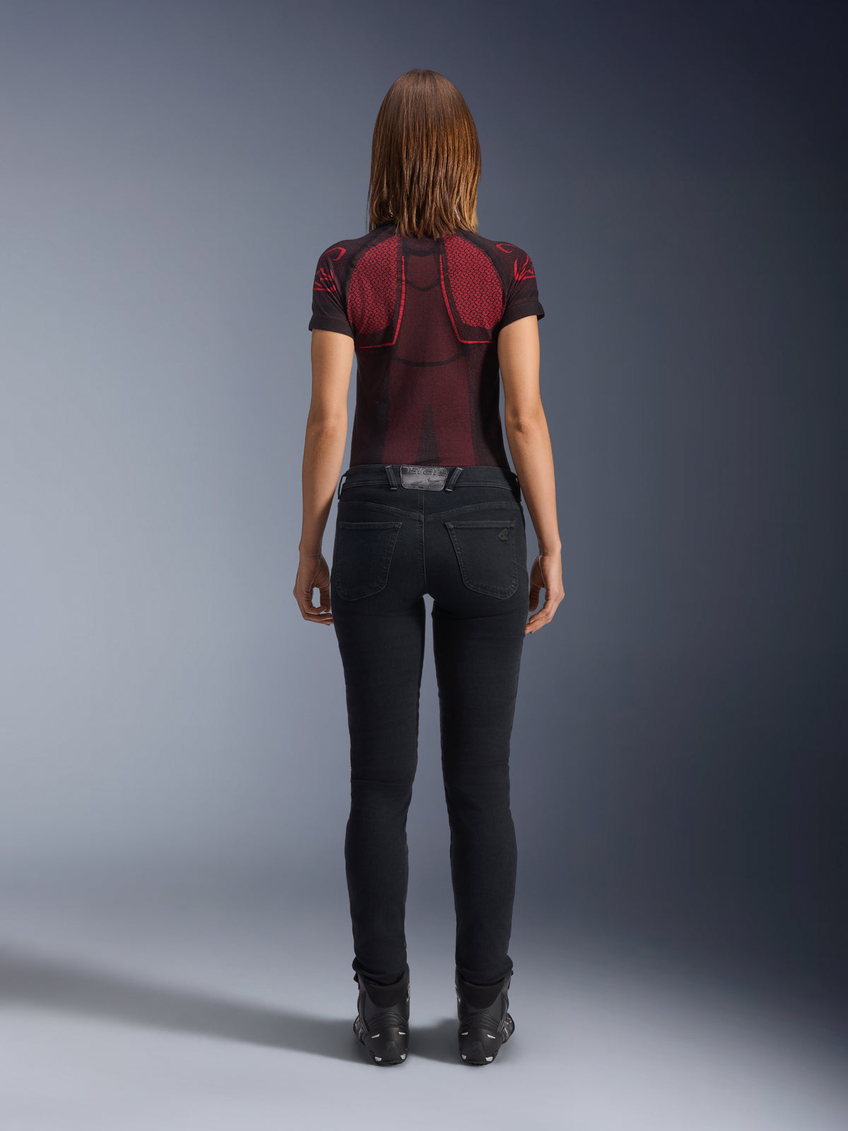 Alpinestars Daisy V3 Women's Riding Denim, preto, calças de motociclismo de corte slim vistas de trás, com design de cintura alta e bolsos traseiros, combinadas com uma camisola técnica com padrão vermelho e preto e botas pretas, equipamento de motociclismo urbano para mulher