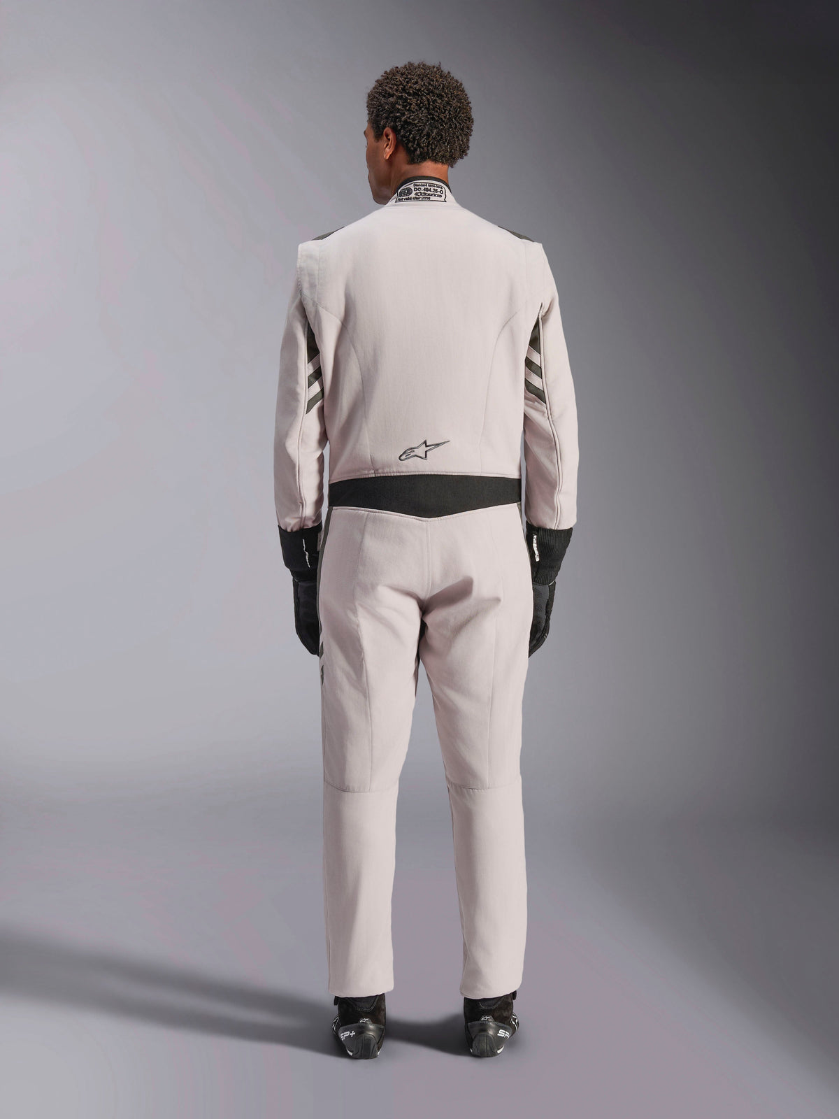 Alpinestars GP Race V3 Suit FIA, Silver Anthracite, vista traseira de fato de automobilismo de uma peça usado com luvas e botas pretas, com painéis elásticos pretos na cintura e nas mangas, design profissional de desporto motorizado