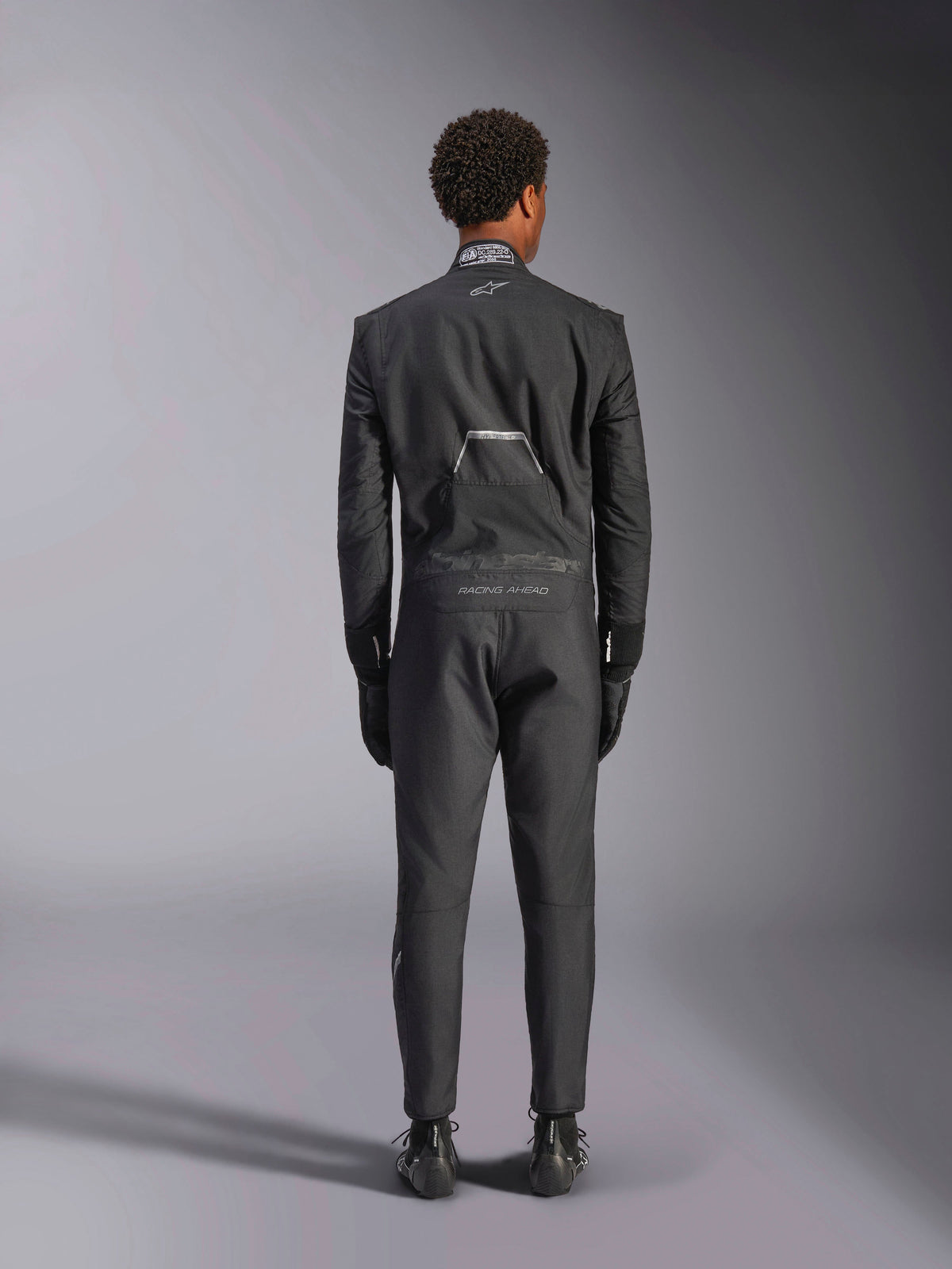 Alpinestars Hypertech V3 Suit, Black Dark Shadow, vista traseira de um fato de automobilismo profissional de peça única, com etiqueta de certificação FIA na gola e logótipos 'Alpinestars' e 'RACING AHEAD' na zona lombar, usado com luvas e botas de corrida pretas a condizer.