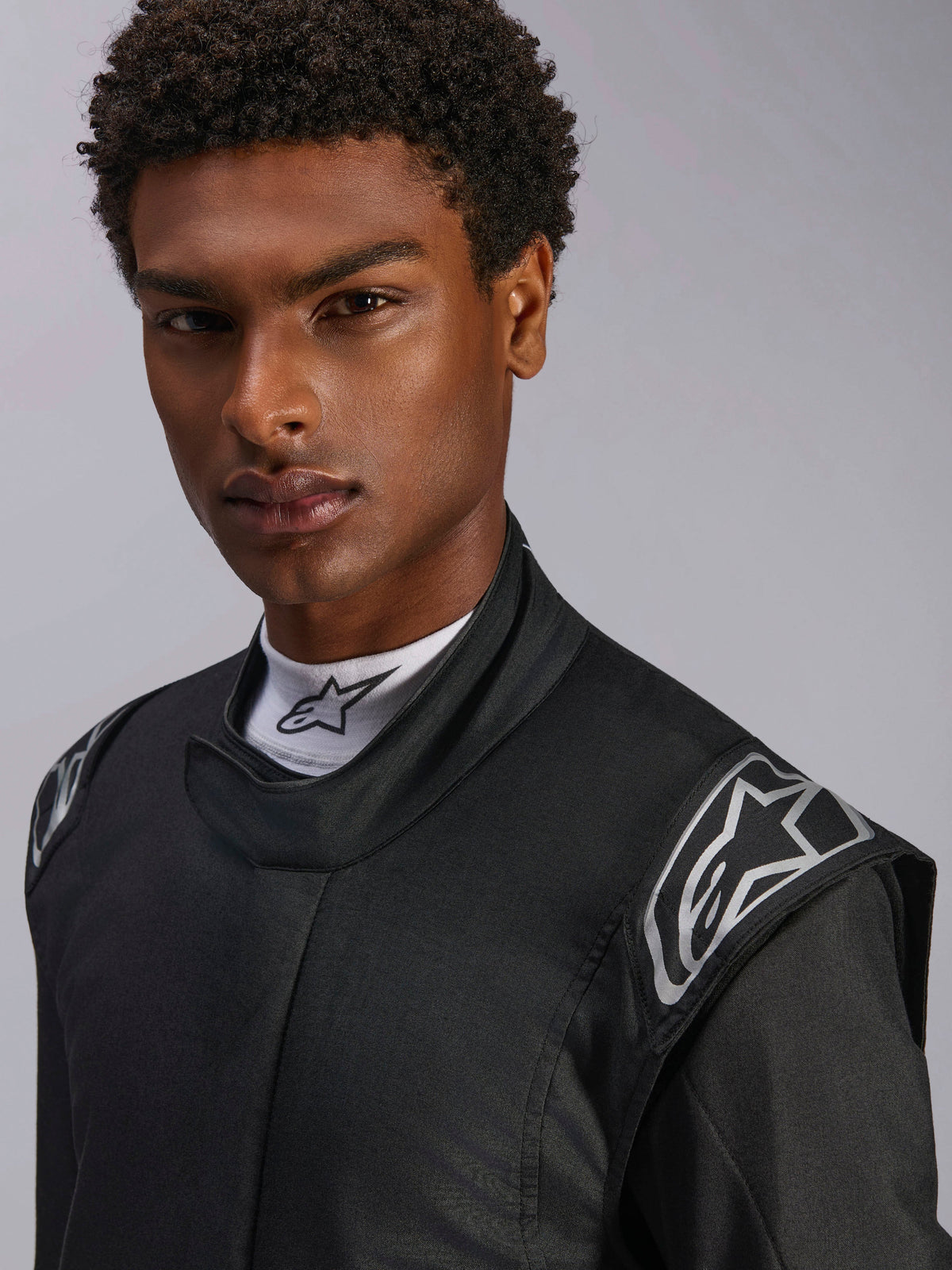 Fato Alpinestars Hypertech V3, fato de automobilismo em Black Dark Shadow, grande plano de modelo masculino, logótipos prateados nos ombros, gola alta com camisola interior Alpinestars branca de gola alta, equipamento de proteção profissional para desportos motorizados