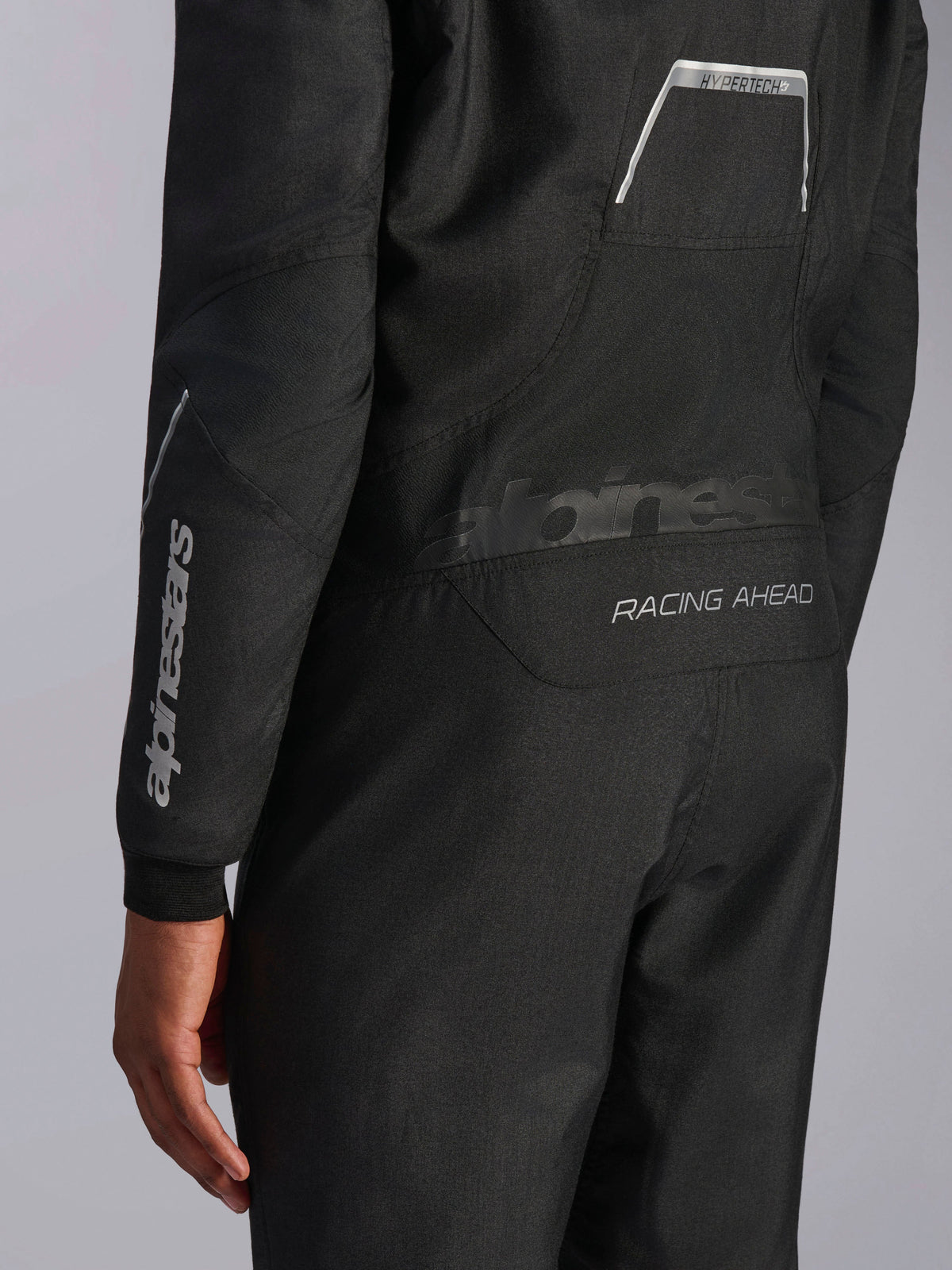 Pormenor do Macacão Alpinestars Hypertech V3 em Preto/Dark Shadow, mostrando os gráficos traseiros 'RACING AHEAD' e 'HYPERTECH V3', com logótipo branco na manga, equipamento profissional de automobilismo.