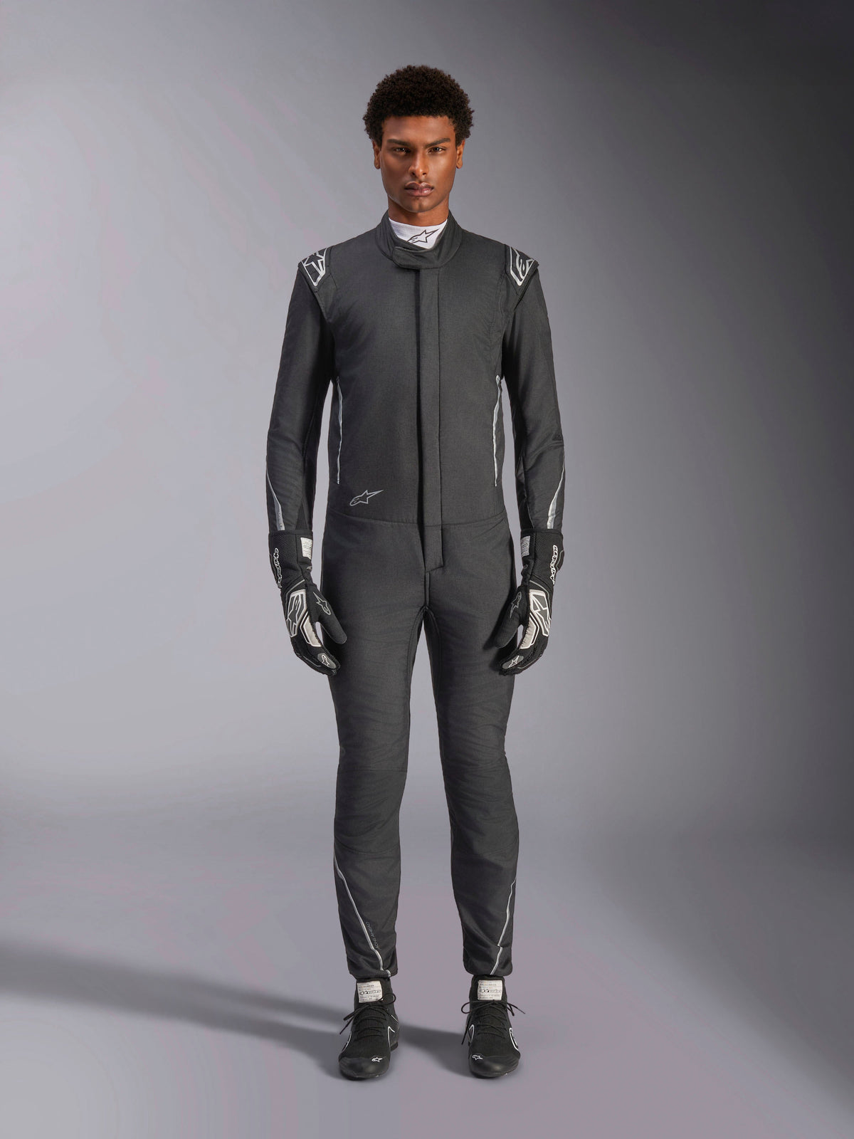 Fato Alpinestars Hypertech V3, fato de automobilismo profissional, Preto e Dark Shadow, vestido por um modelo com luvas e sapatos de corrida a condizer, corte atlético ajustado com detalhes prateados e logótipos da marca, equipamento de performance para desportos motorizados