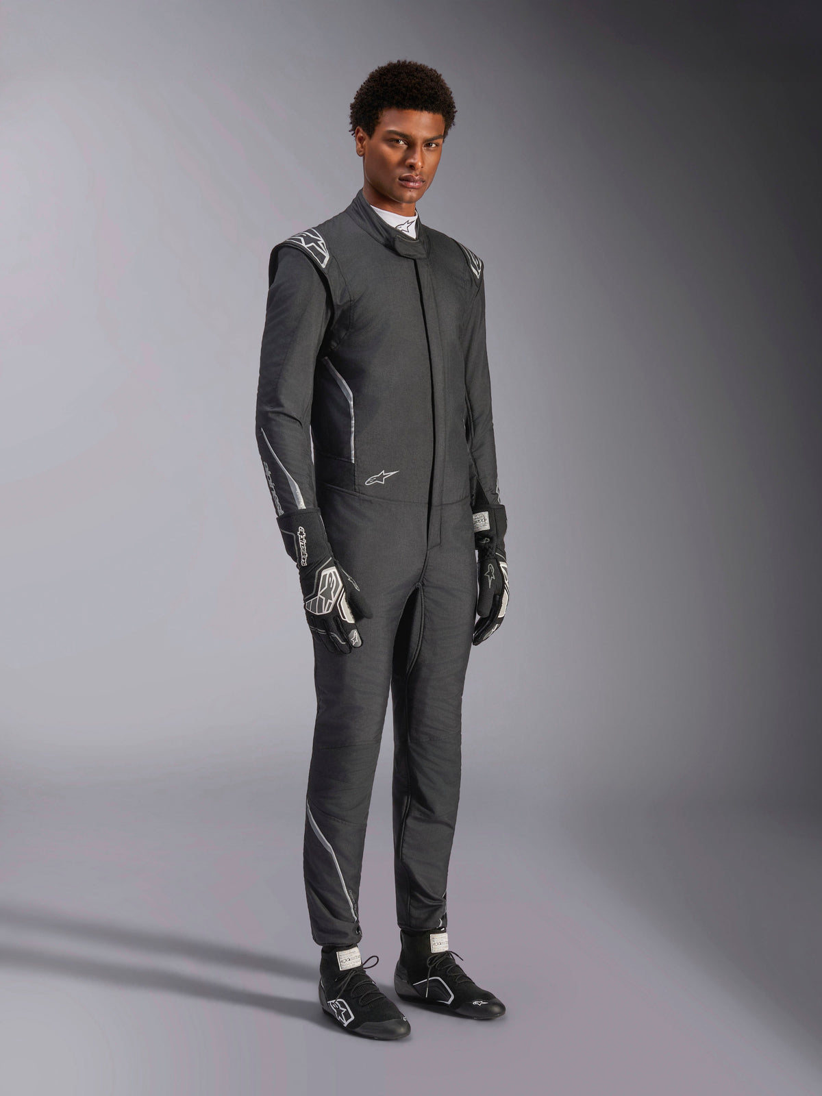 Fato Alpinestars Hypertech V3, fato de automobilismo, preto e dark shadow, usado por modelo masculino com luvas pretas e sapatos de corrida a condizer, corte atlético slim fit, com logótipos Alpinestars e detalhes em cinzento claro, equipamento de proteção profissional para automobilismo