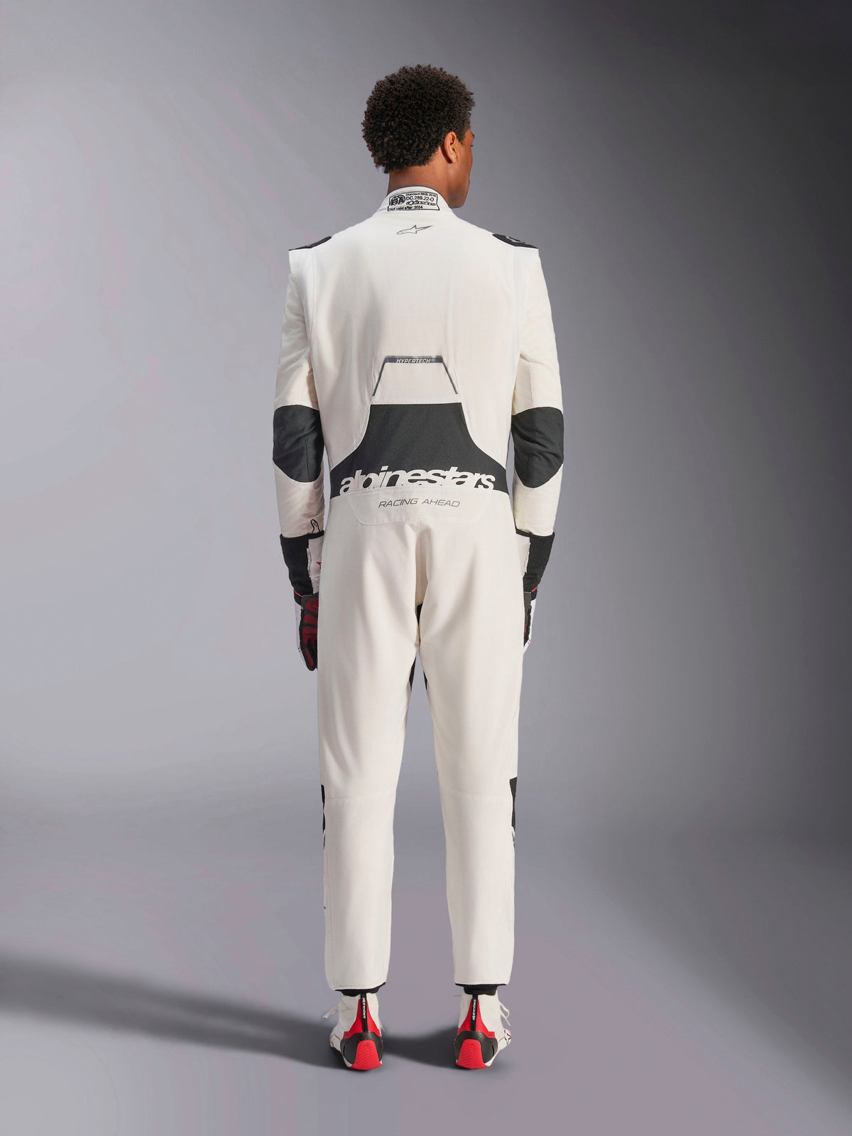Vista traseira de um modelo a usar o Macacão Alpinestars Hypertech V3 em Branco, Preto e Prata, destacando o grande painel elástico lombar preto com as marcas 'alpinestars' e 'RACING AHEAD', inserções pretas nos cotovelos, etiqueta de certificação FIA na gola e um corte atlético concebido para competição profissional.