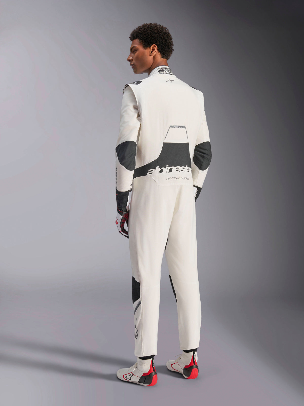 Fato Alpinestars Hypertech V3, fato de automobilismo profissional, branco, preto e prateado, vista traseira do modelo mostrando painéis elásticos pretos na zona lombar e cotovelos, marca Alpinestars nas costas, combinado com luvas e botas de competição a condizer para um visual de desporto motorizado completo
