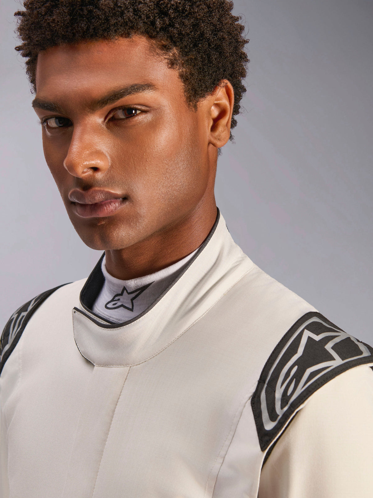 Close-up de um modelo masculino a usar o fato Alpinestars Hypertech V3 em Branco, Preto e Prata, com gola alta de fecho seguro e logótipos Alpinestars nos ombros, equipamento profissional de automobilismo