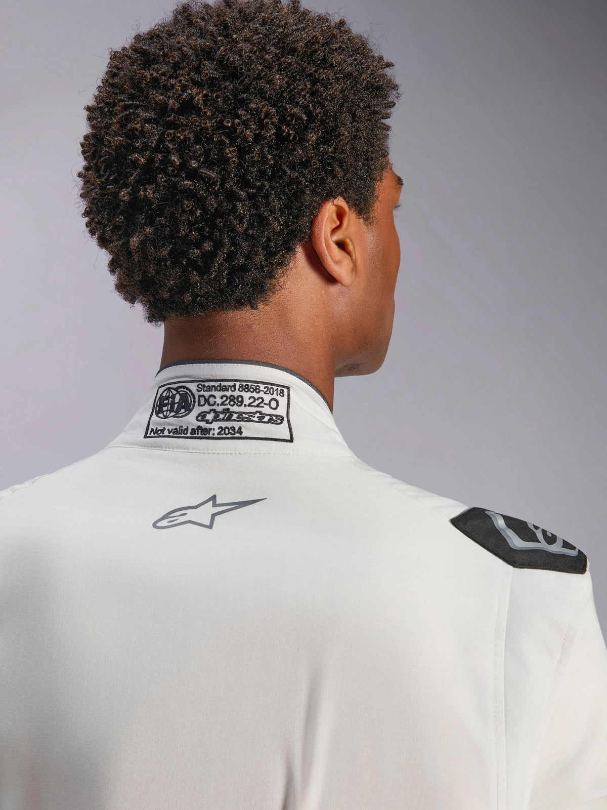 Fato Alpinestars Hypertech V3, branco, vista de detalhe da gola traseira e do ombro, com etiqueta de certificação FIA 8856-2018, logótipo Alpinestars cinzento impresso e pega de extração preta, equipamento de corrida profissional