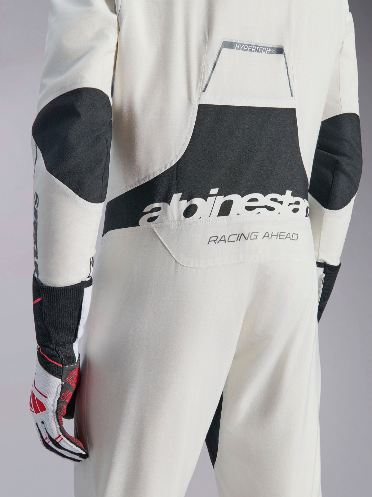Macacão Alpinestars Hypertech V3, macacão de automobilismo em branco, preto e prateado, vista traseira com grande logótipo 'alpinestars' preto e texto 'RACING AHEAD', painéis elásticos pretos nos cotovelos e zona lombar, vestuário de proteção profissional para automobilismo.