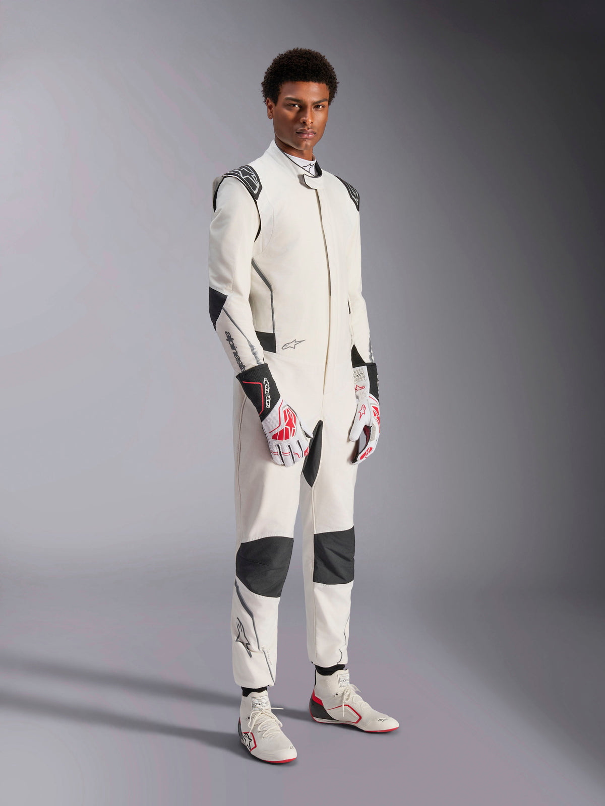 Modelo masculino a usar o Macacão Alpinestars Hypertech V3, em branco com detalhes em preto e prateado, equipamento profissional de automobilismo com painéis pretos nos ombros e joelhos, complementado por luvas de corrida brancas e vermelhas e botas de corrida brancas
