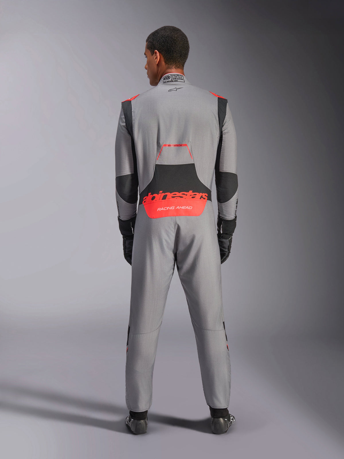 Fato Alpinestars Hypertech V3, fato de automobilismo profissional, cinzento médio com detalhes em preto e vermelho fluo, vista traseira num modelo com marca Alpinestars a vermelho na zona lombar, construção leve em Nomex para performance em pista