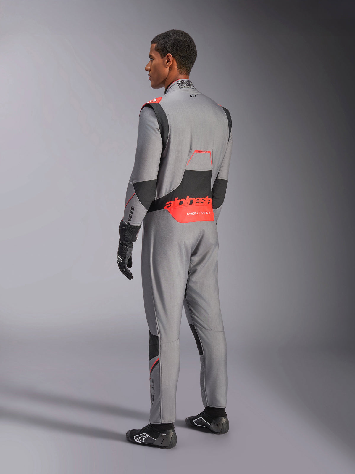 Macacão Alpinestars Hypertech V3 em cinzento médio, preto e vermelho fluo, vista traseira de três quartos, com logótipo Alpinestars vermelho na zona lombar, painéis elásticos pretos e ajuste atlético para automobilismo profissional