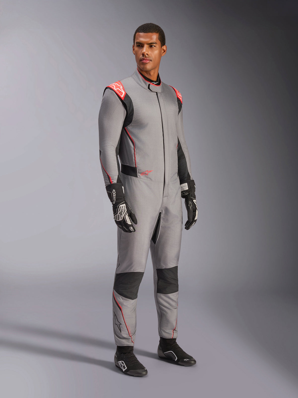 Modelo masculino a usar o fato Alpinestars Hypertech V3 em cinzento médio com pormenores em preto e vermelho fluo, fato de automobilismo profissional combinado com luvas e sapatos de corrida pretos, com uma construção leve em Nomex de duas camadas, pormenores vermelhos nos ombros e um ajuste atlético