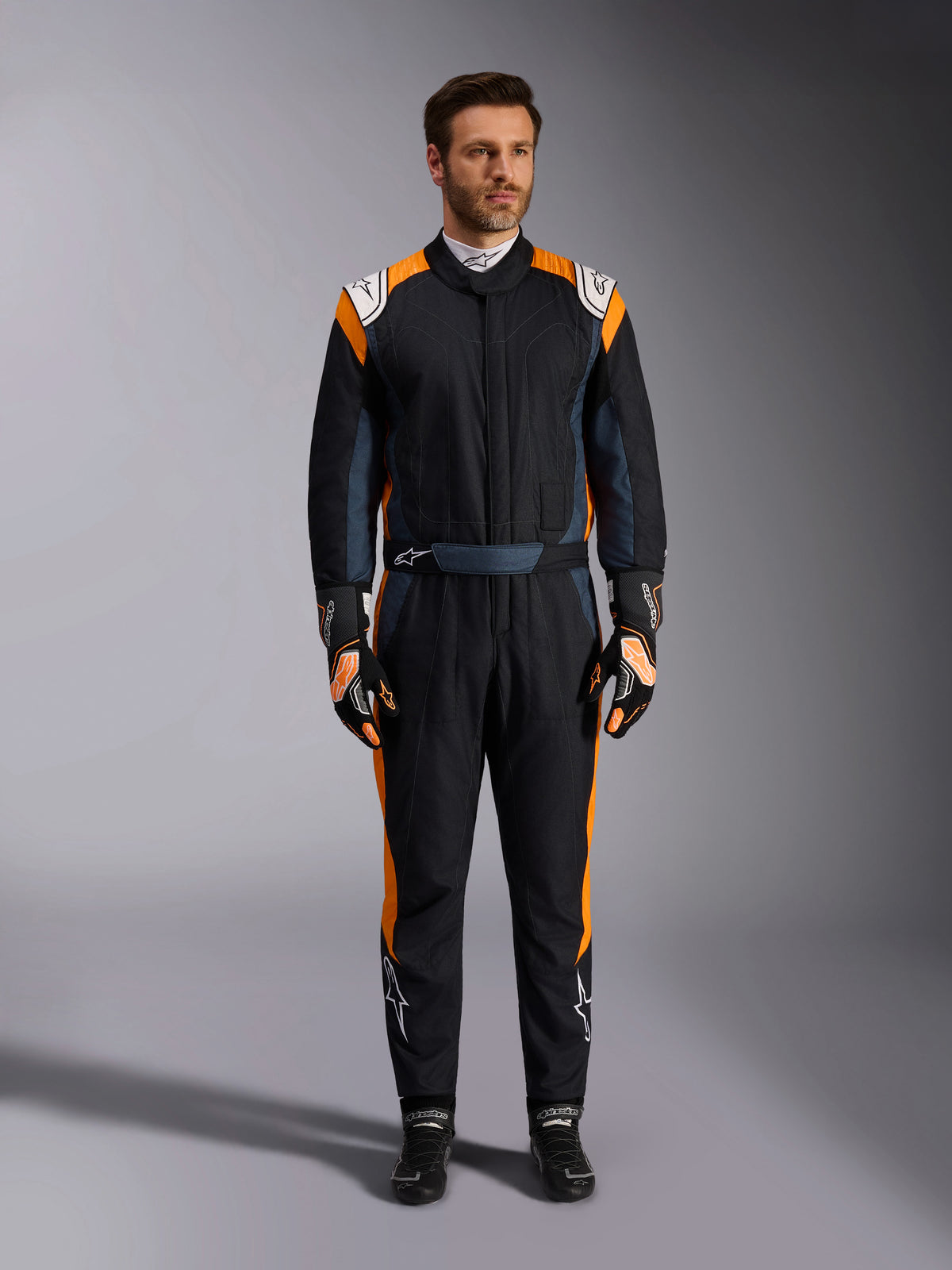Alpinestars GP Pro Comp V2 FIA Suit, fato de automobilismo em Black Asphalt e Laranja Fluo, modelo masculino a usar equipamento de proteção completo com luvas e sapatos de corrida a condizer, inclui logótipos Alpinestars brancos e painéis laterais em laranja vibrante, vestuário profissional de desportos motorizados