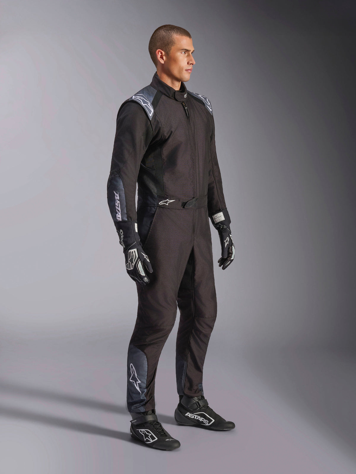 Macacão Alpinestars KMX-5 V3, fato de karting em Black Dark Shadow, usado com luvas e botas de corrida pretas a condizer, com perfurações cortadas a laser e painéis de malha tecnológica, design elegante de camada dupla com logótipos brancos para desporto motorizado profissional