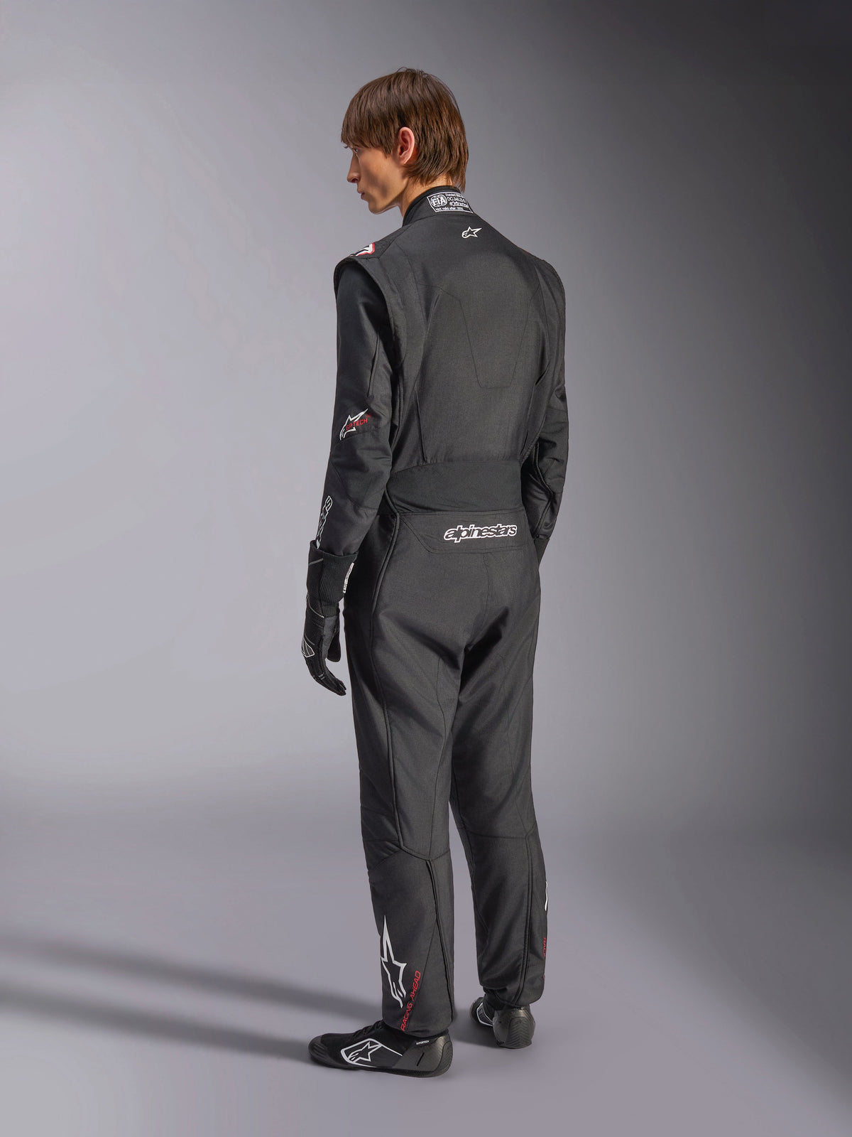Alpinestars GP Tech V4 Suit, Auto Racing Suit, Black with white accents, back view of model wearing the suit with racing gloves and shoes, featuring white branding on the lower back and leg, professional motorsport safety gear