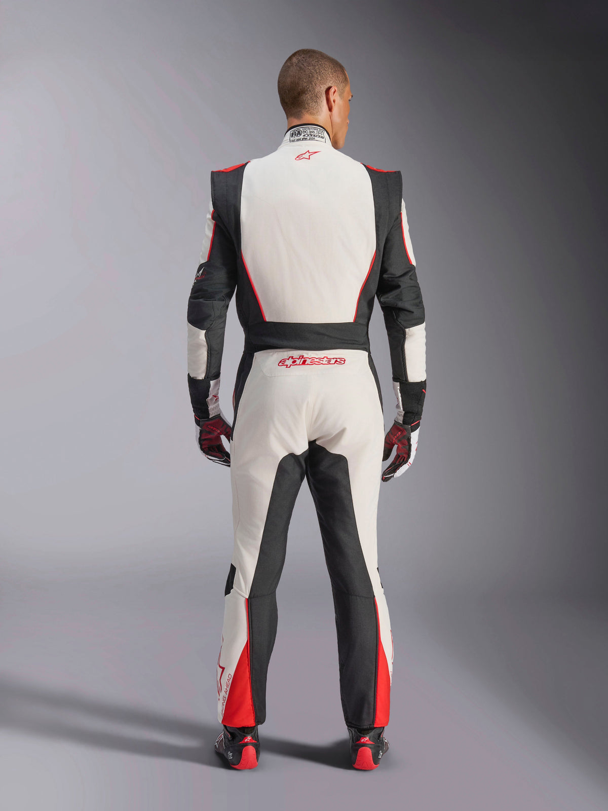 Alpinestars GP Tech V4 Suit, macacão de automobilismo, branco, preto e vermelho, vista traseira do piloto a usar o fato de peça única, com painéis elásticos pretos nas laterais e ombros, detalhes em vermelho na parte inferior das pernas, logótipo Alpinestars na zona lombar e na gola, equipamento de automobilismo profissional com certificação FIA