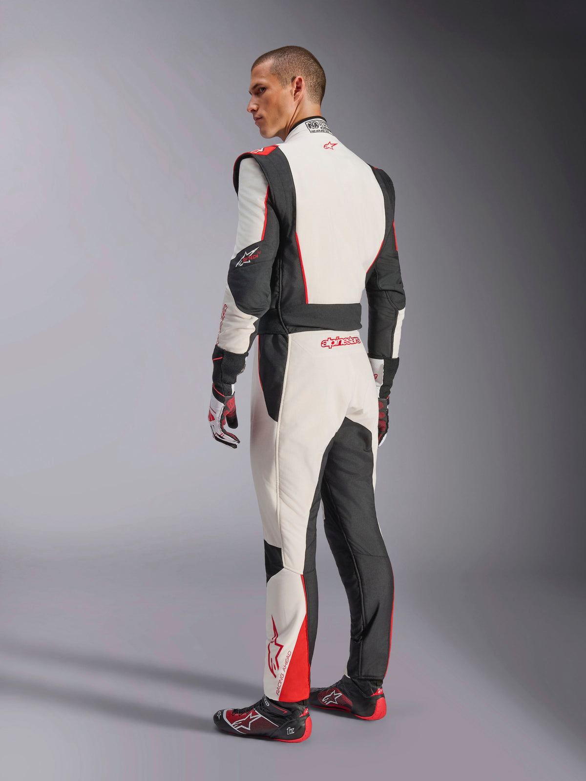 Macacão Alpinestars GP Tech V4, macacão de competição em branco, preto e vermelho, vista traseira com painéis elásticos ergonómicos na zona lombar e pernas, combinado com luvas e sapatos de corrida a condizer, vestuário profissional de automobilismo.