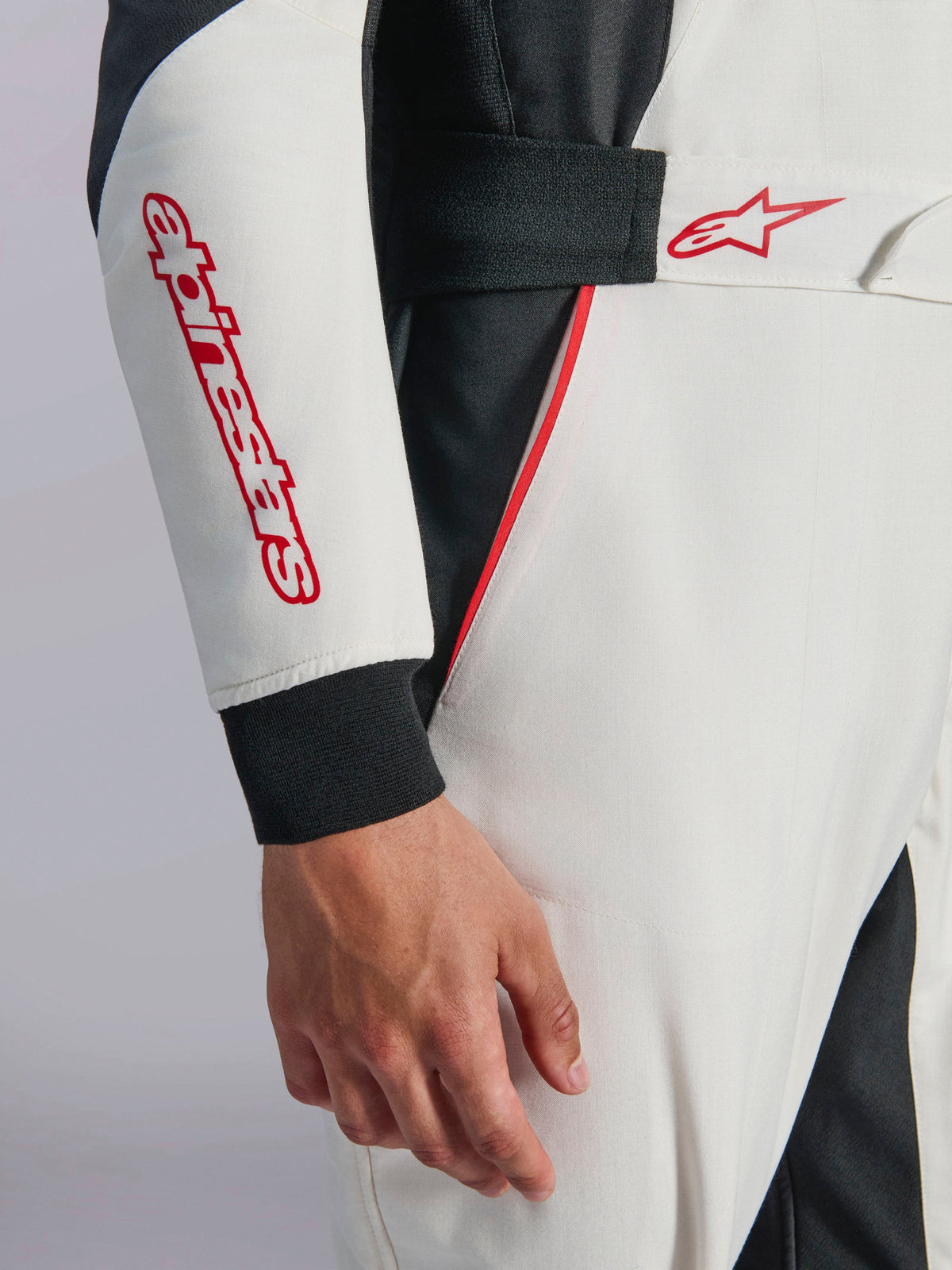 Macacão Alpinestars GP Tech V4, macacão de automobilismo branco, preto e vermelho, grande plano da manga e da cintura, com marca e logótipo de estrela Alpinestars em vermelho, cintura elástica com fecho de gancho e laço, e debrum vermelho sobre tecido técnico branco e preto