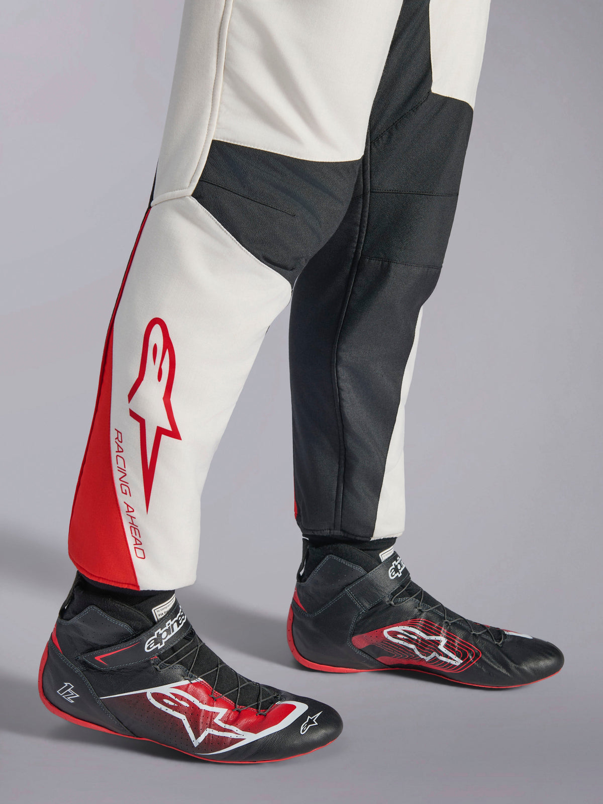 Macacão Alpinestars GP Tech V4, macacão de automobilismo, branco, preto e vermelho, vista da parte inferior da perna com painéis brancos e pretos e detalhes vermelhos, marca 'RACING AHEAD', usado com sapatilhas de competição pretas, equipamento profissional de desportos motorizados