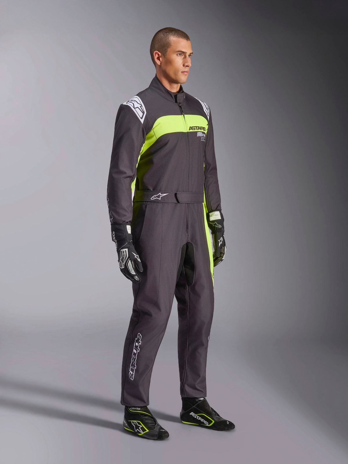 Modelo masculino com o Macacão Alpinestars KMX-9 V3 Graphic 3 em preto e amarelo néon, equipamento profissional de karting com painéis vibrantes e detalhes de logótipo em branco, acompanhado por luvas e sapatos de corrida coordenados, num fundo de estúdio.