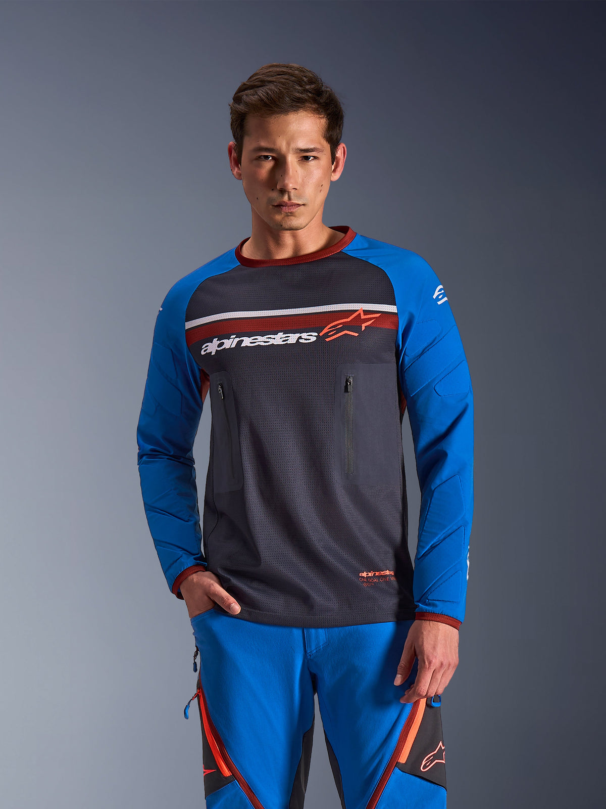 Man wearing Alpinestars Maxdura Dual Jersey in Deep Gray and Royal Blue with orange accents, featuring a full mesh torso and zippered chest pockets, paired with matching blue adventure riding pants, technical dual-sport and off-road apparel