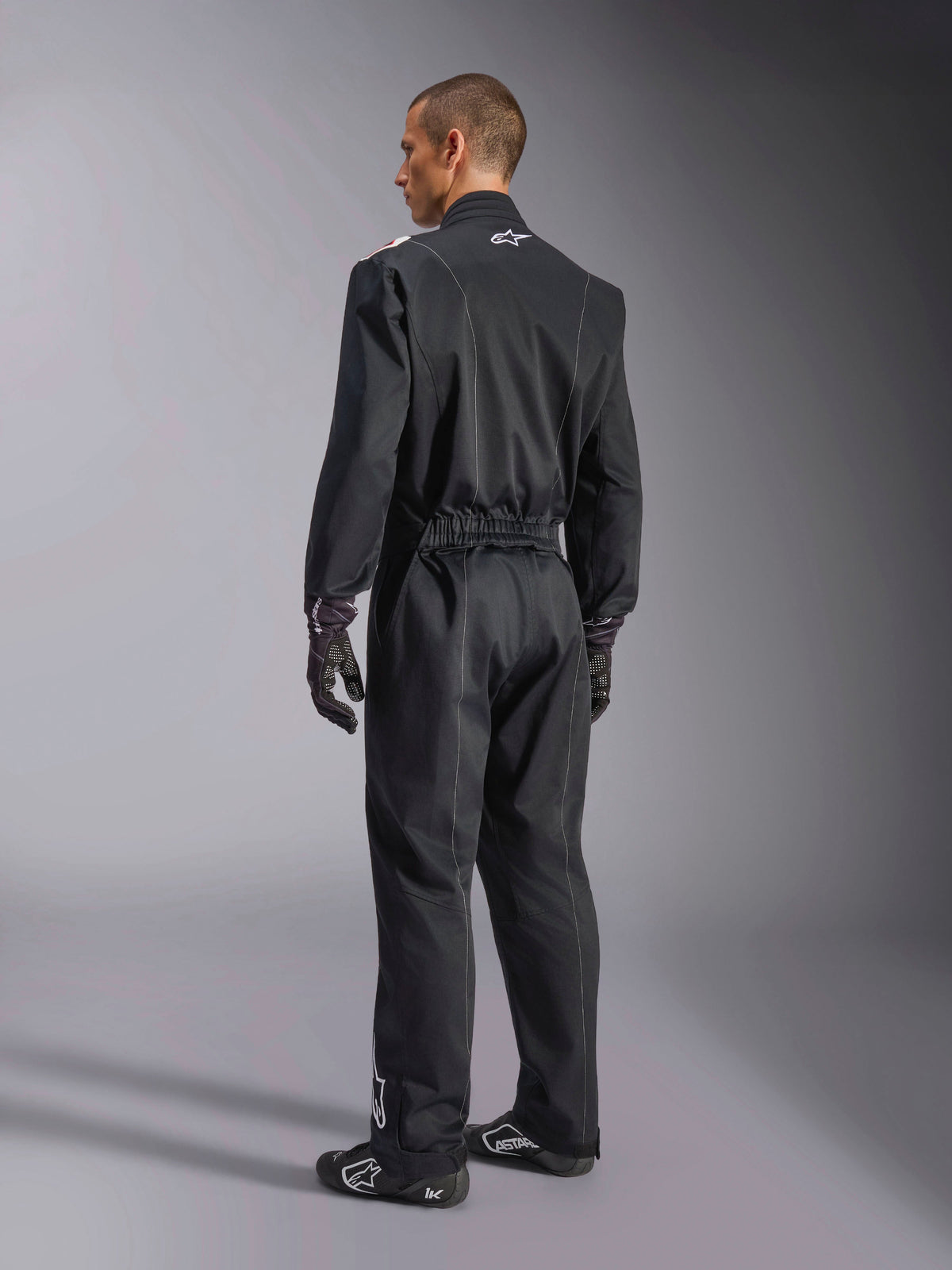 Alpinestars Kart Indoor V2 Suit, fato de karting de peça única em preto visto de um ângulo de três quartos de costas, com cintura elástica e logótipo branco no pescoço, usado com luvas e sapatos de corrida pretos, design durável para karting recreativo